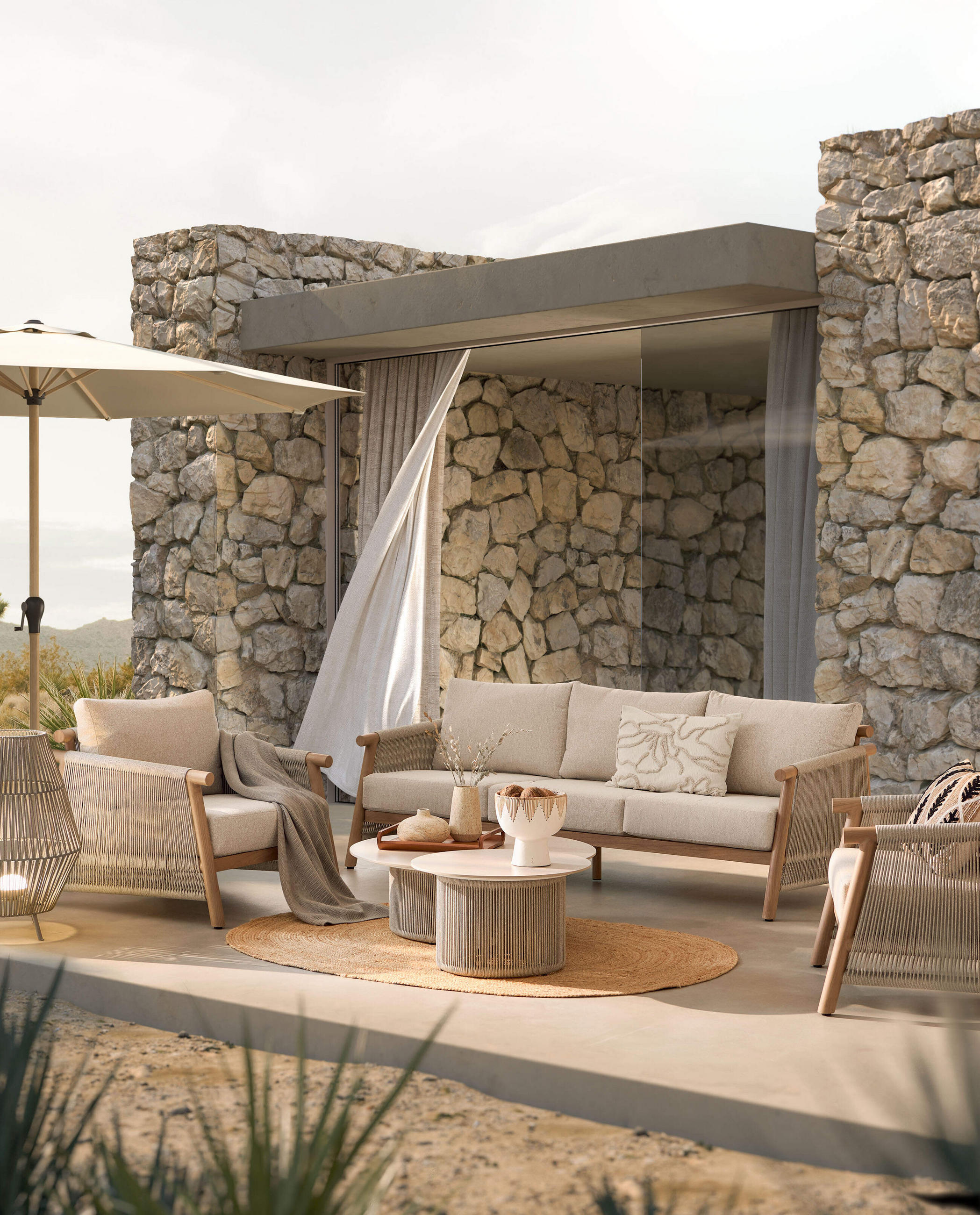 Outdoor-Sitzgruppe in Beige mit Kissen und Rundteppich, vor Natursteinwand mit Sonnenschirm und Vorhang