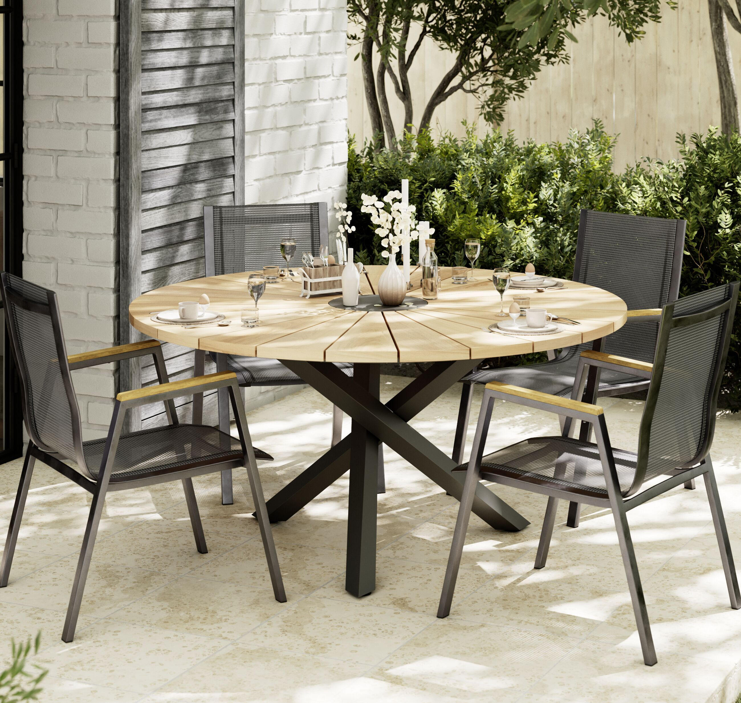 Runder Holzgartentisch mit vier grauen Stühlen auf einer Terrasse, gedeckt mit Geschirr und Vasen, Hecke im Hintergrund.