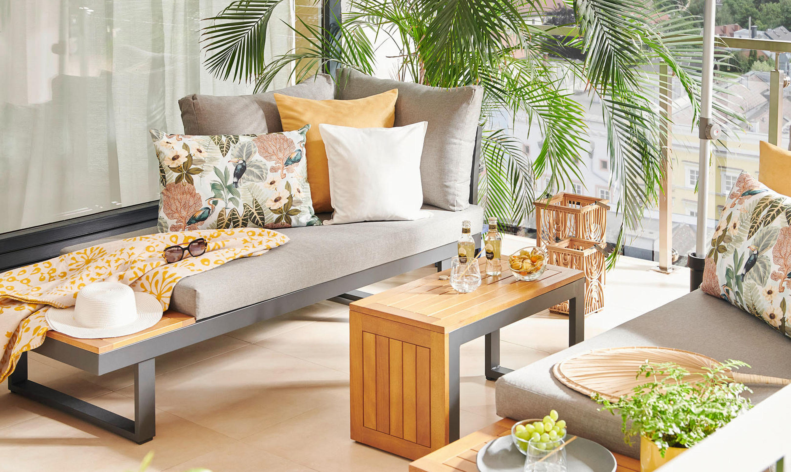 Gemütliche Outdoor-Lounge mit grauen Polstern, Holztisch, Kissen und Palmen auf einem sonnigen Balkon