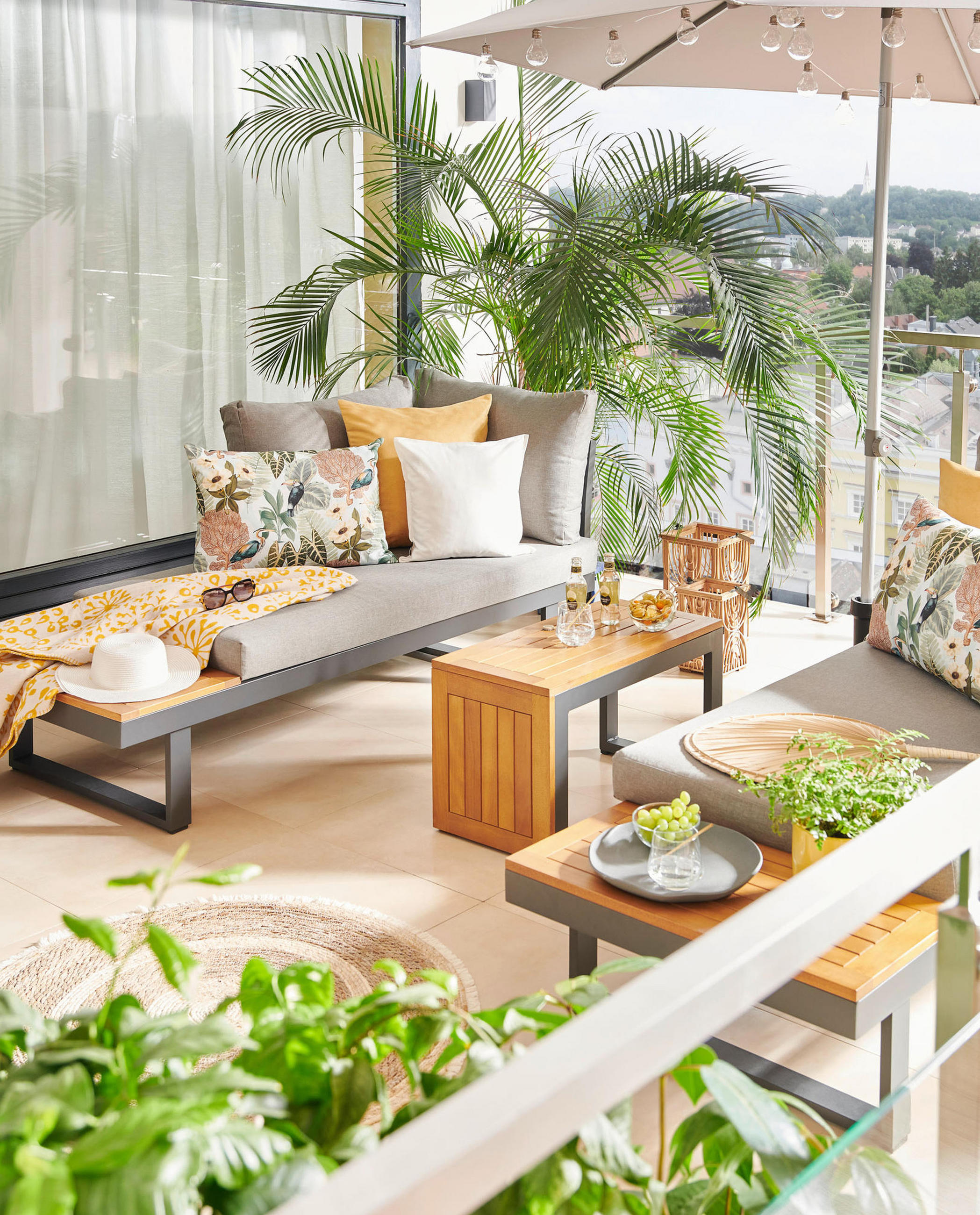 Gemütliche Outdoor-Lounge mit grauen Polstern, Holztisch, Kissen und Palmen auf einem sonnigen Balkon