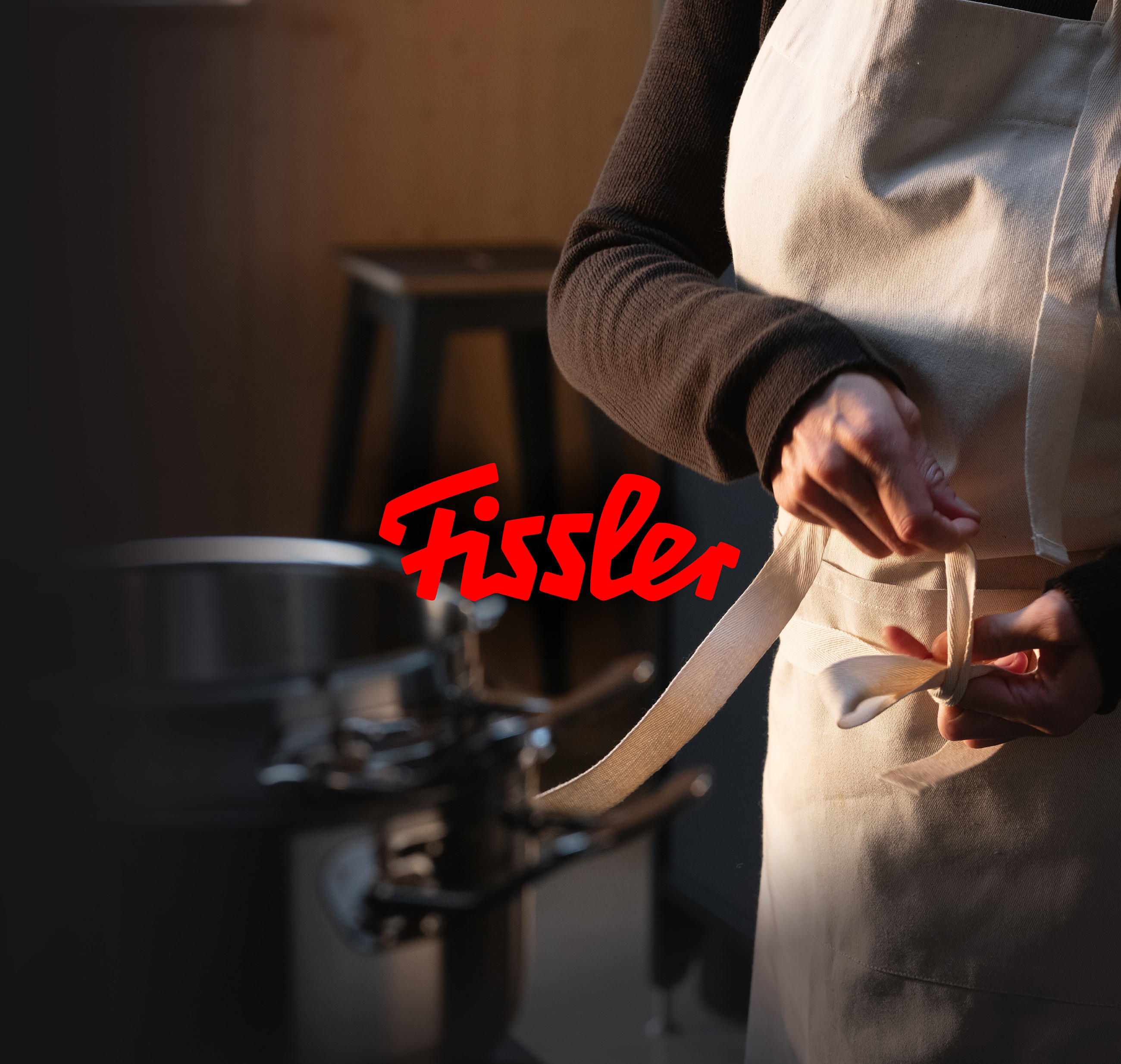 Person bindet die Schürze in einer Küche; im Vordergrund ein glänzender Topf. Zentral platziert ist das rote FISSLER‑Markenlogo.
