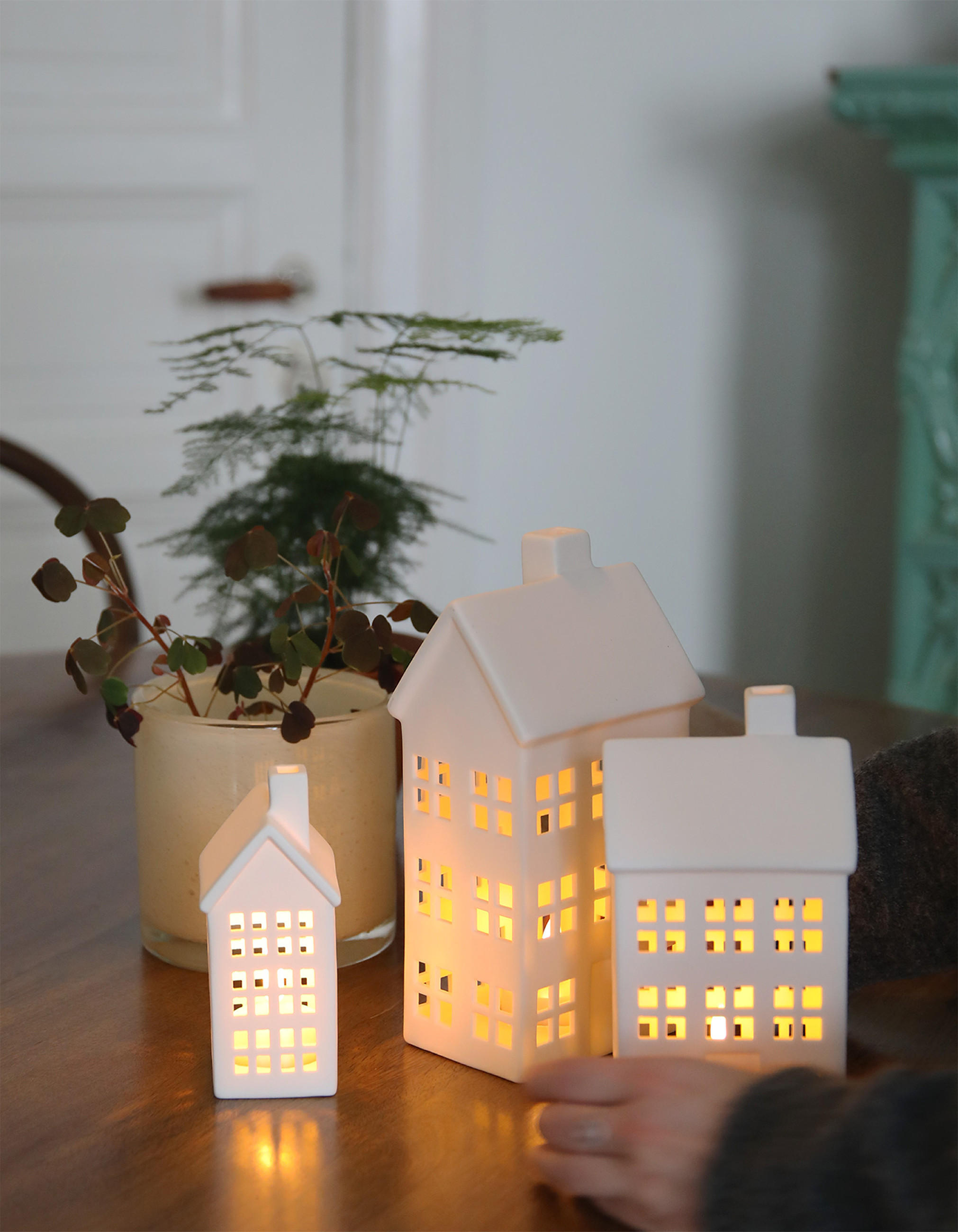 Leuchtende MARKSLÖJD Dekoleuchten in Hausform aus weißer Keramik mit warmem Licht auf einem Holztisch neben einer Vase mit Grünzweigen.