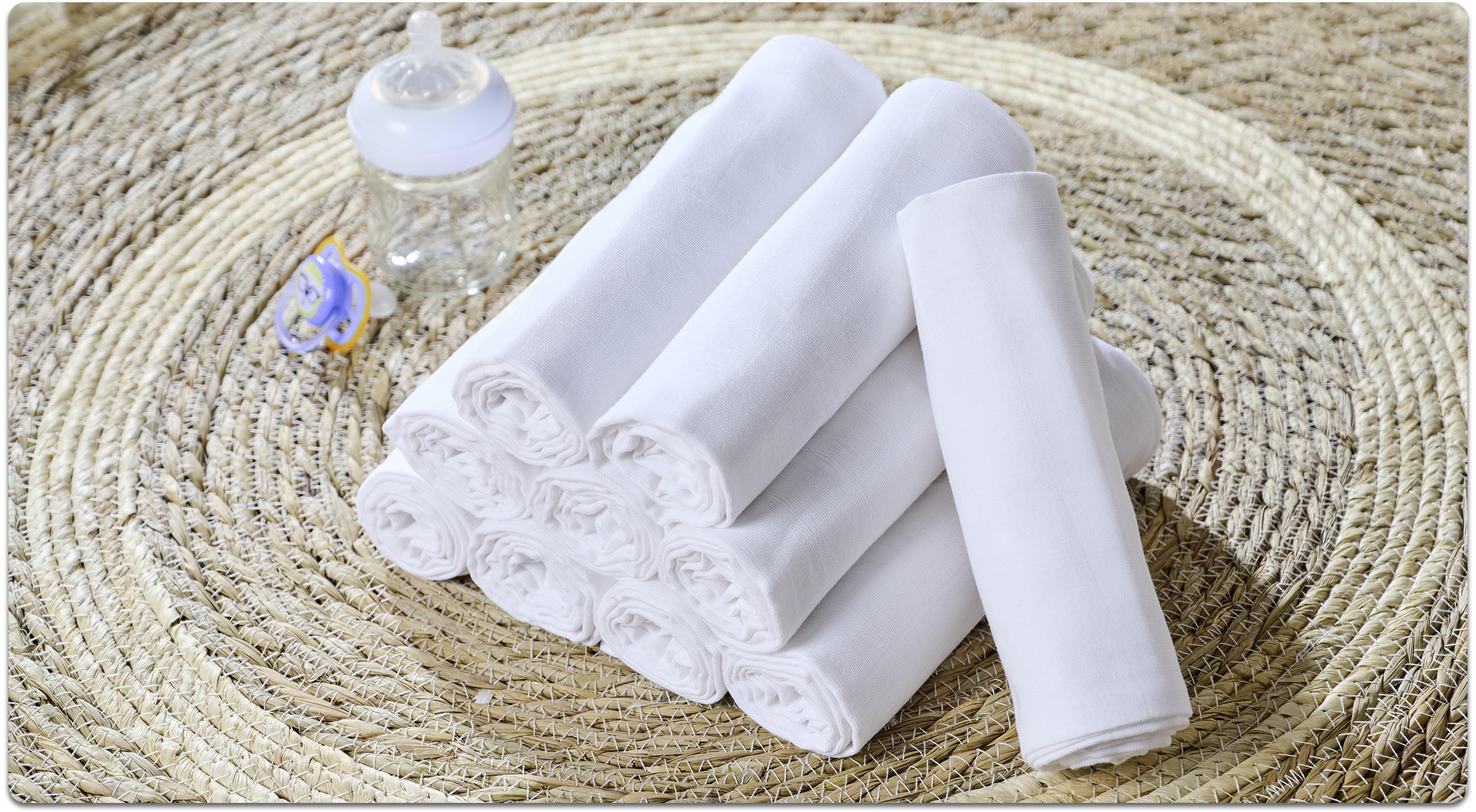Mehrere weiße Stoffwindeln mit Spitzenkante liegen gerollt auf einem runden Juteteppich, daneben Babyflasche und Schnuller.