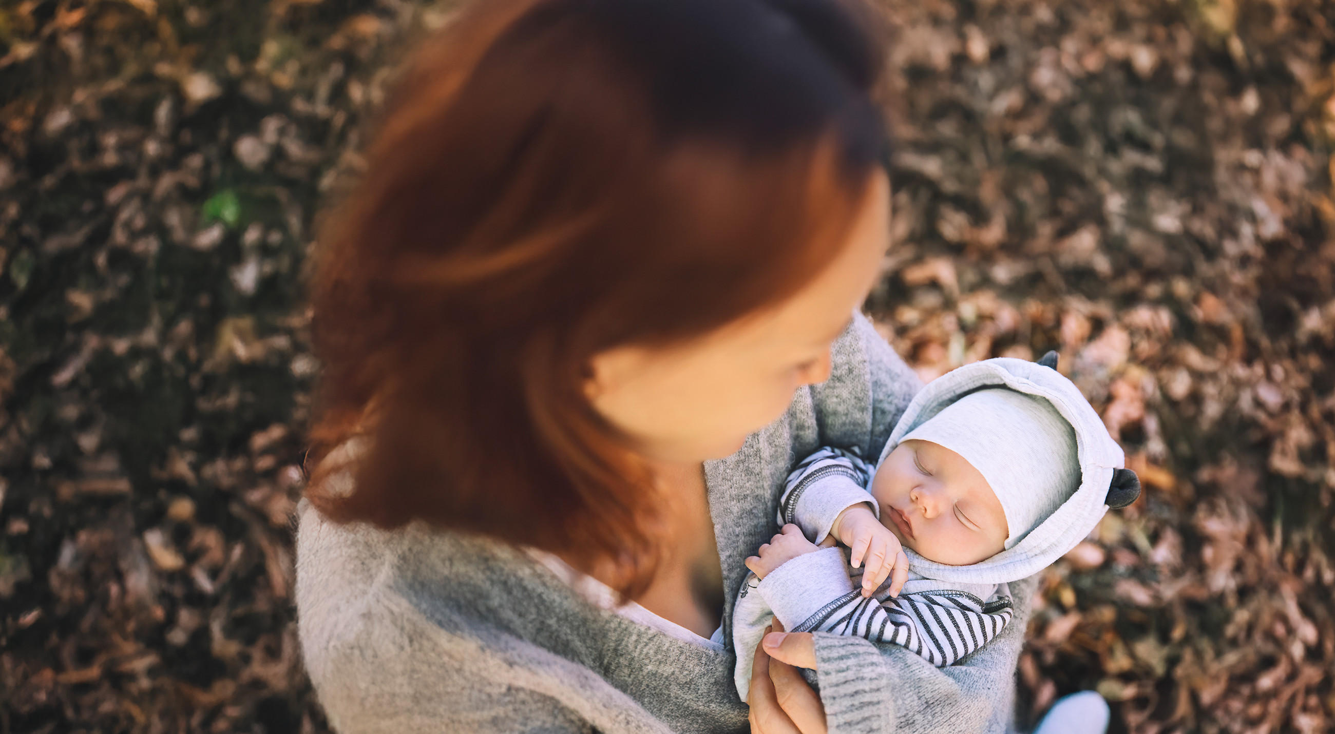 Eine Person hält ein schlafendes Baby mit Mütze und gestreiftem Overall im Arm, umgeben von herbstlichem Laub.