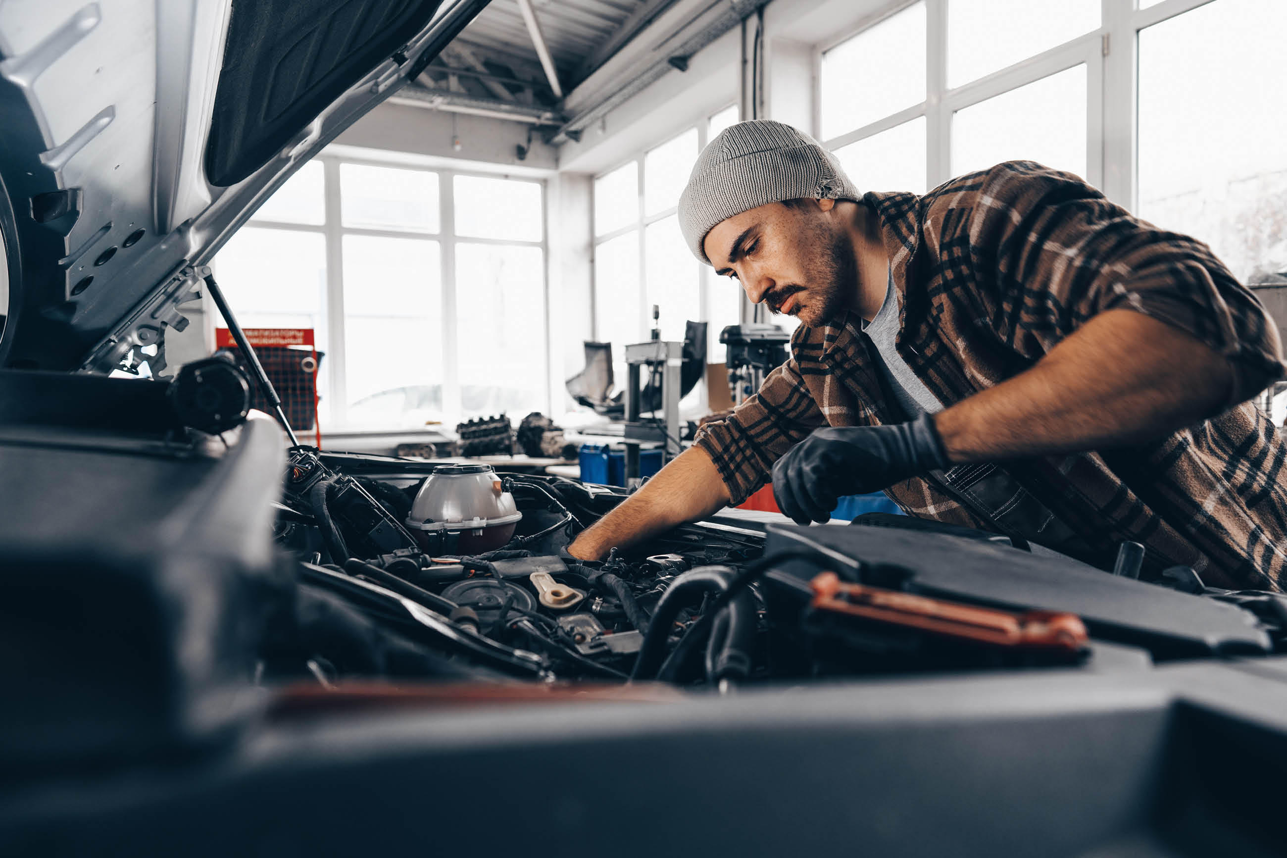 Mann in einem Werkstatt mit Mütze und Handschuhen, der an einem Auto arbeitet und Reparaturen durchführt.