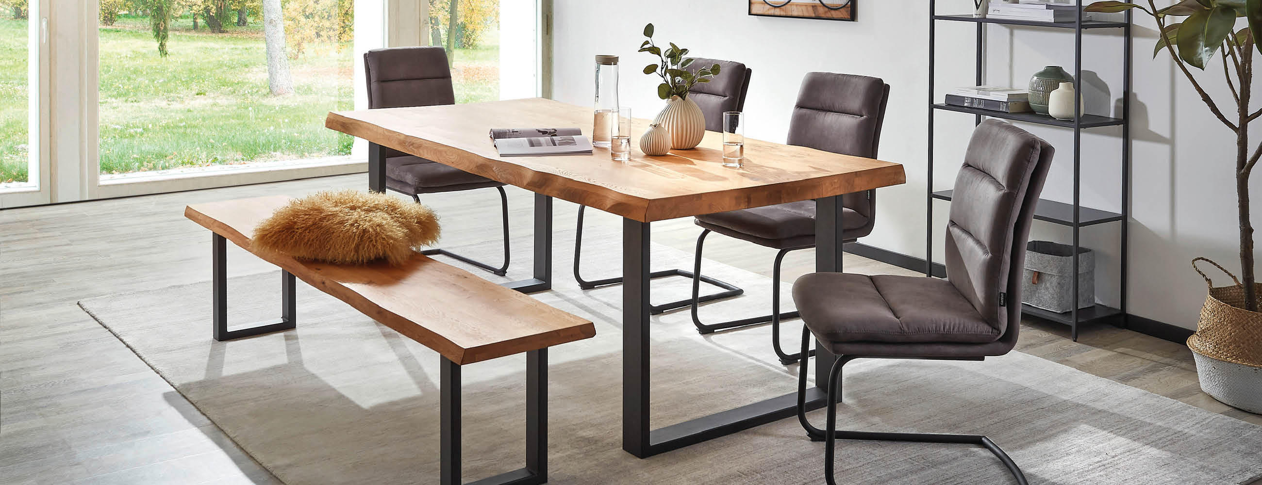 Esszimmer mit rechteckigem Holztisch auf schwarzen Metallbeinen, zwei gepolsterten Stühlen mit Rückenlehne und einer Holzbank mit Fellkissen, großer Fensterfront im Hintergrund