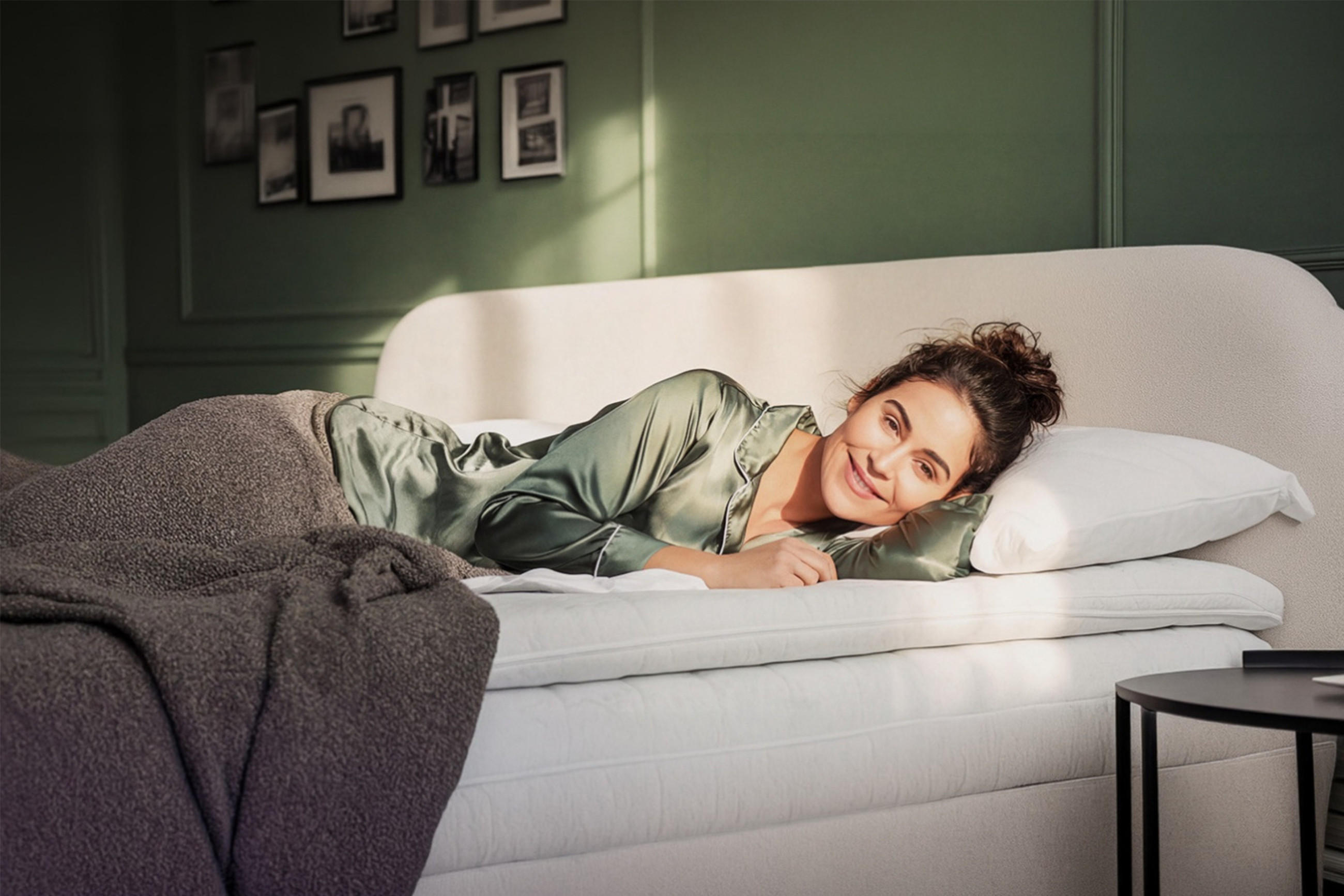 Lächelnde Frau in grün schimmerndem Satin-Pyjama liegt entspannt auf einem weißen Bett mit Topper und Kissen, eingehüllt in eine graue Decke, vor einer grünen Wand mit Bilderrahmen.