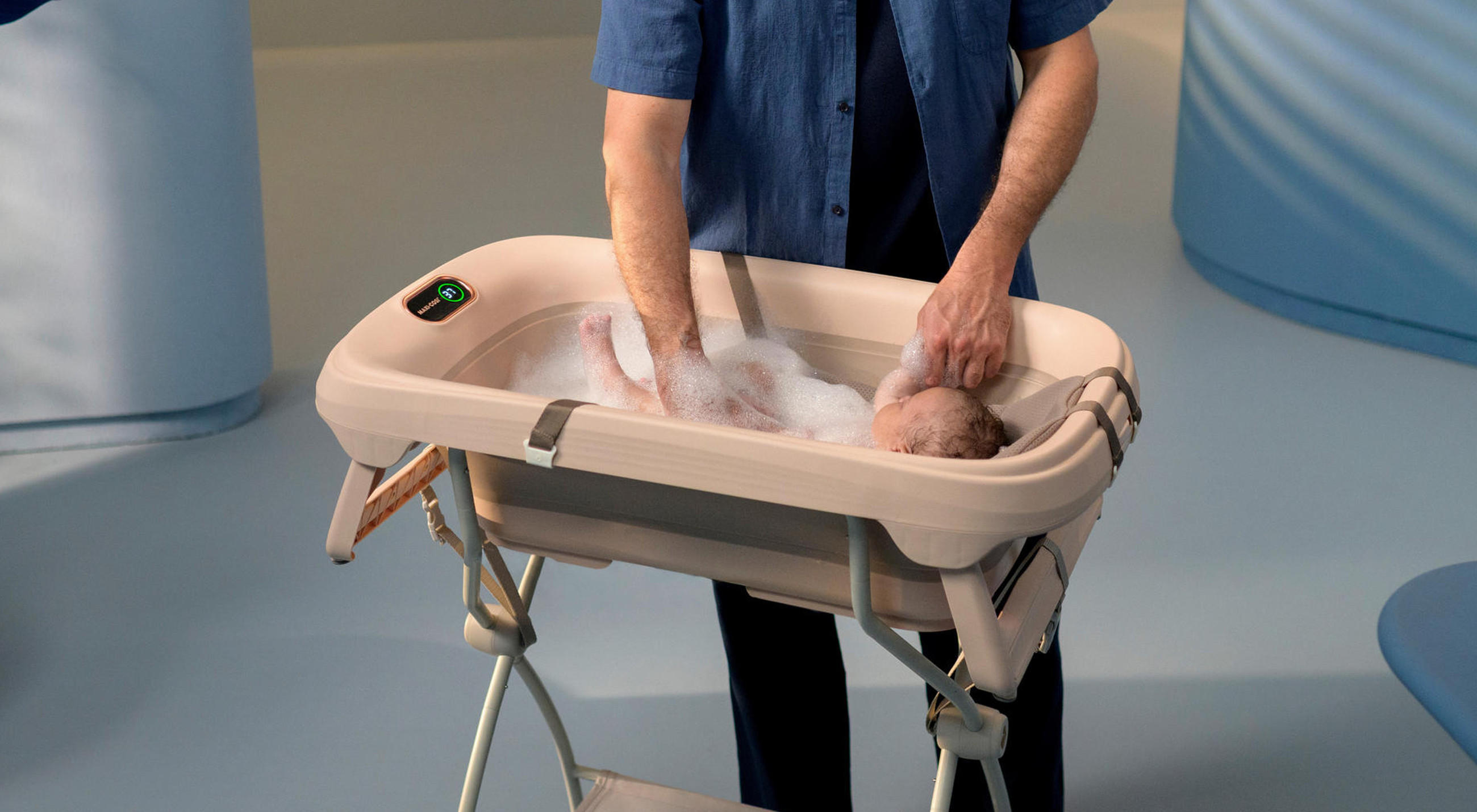 Ein Baby wird in einer beigefarbenen Babybadewanne auf einem Klappständer gebadet, während eine erwachsene Person es vorsichtig hält und wäscht; die Wanne verfügt über ein digitales Bedienfeld