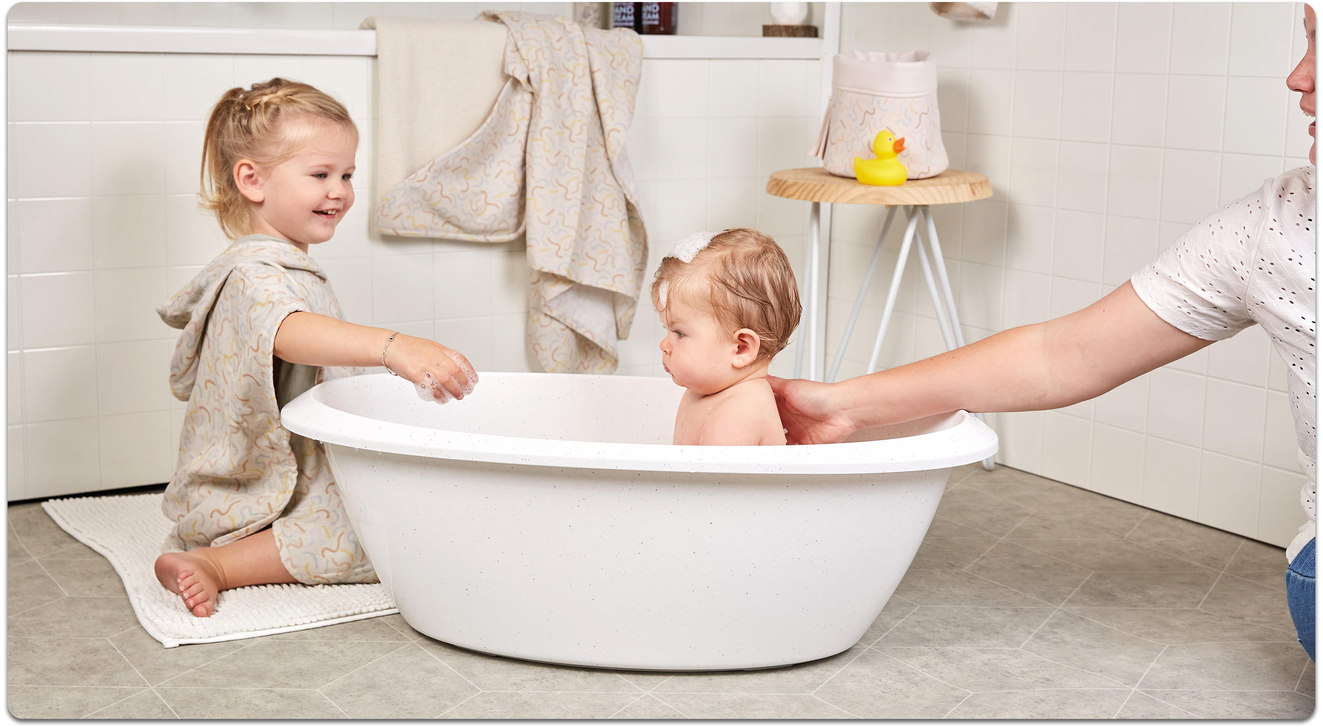 Zwei Kleinkinder baden in einer weißen Babywanne im Badezimmer; rechts reicht eine erwachsene Person den Arm ins Bild, im Hintergrund hängen Handtücher.