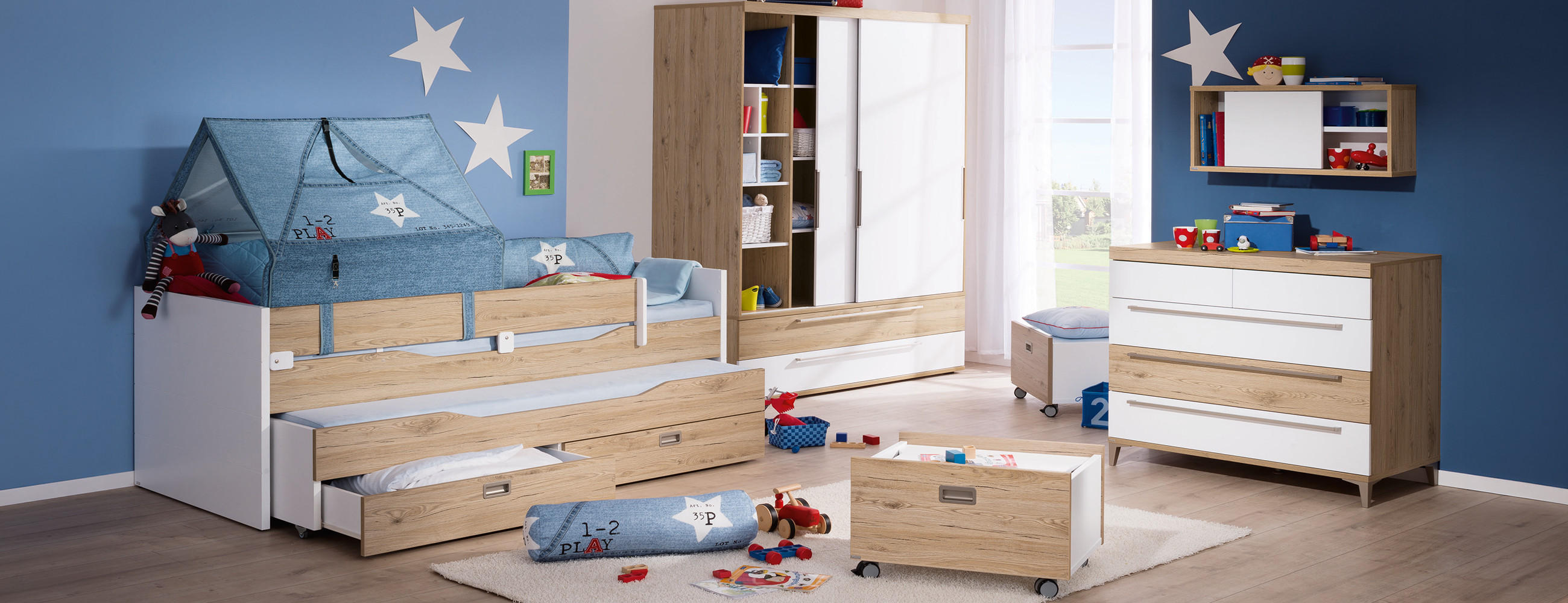 Kinderzimmer mit blau gestrichener Wand, einem Bett mit Netz, Kommode, Regal und Spielzeug auf dem Boden.