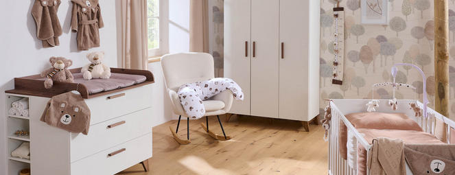 Helles Babyzimmer mit Wickelkommode und gepolsterter Auflage, Teddys und Bademänteln an der Wand, beigem Schrank, Schaukelstuhl mit Stillkissen sowie Babybett mit Mobile vor gemusterter Tapete.