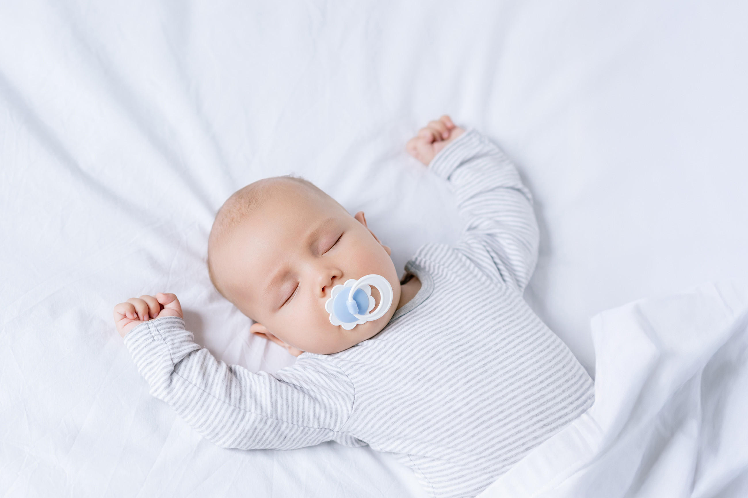 Ein Baby liegt auf dem Rücken auf einer weißen Bettdecke, die Arme entspannt nach oben ausgestreckt. Es trägt einen langärmligen, hellgrau gestreiften Strampler, und die Szene wirkt ruhig und weich durch das helle, gleichmäßige Licht.