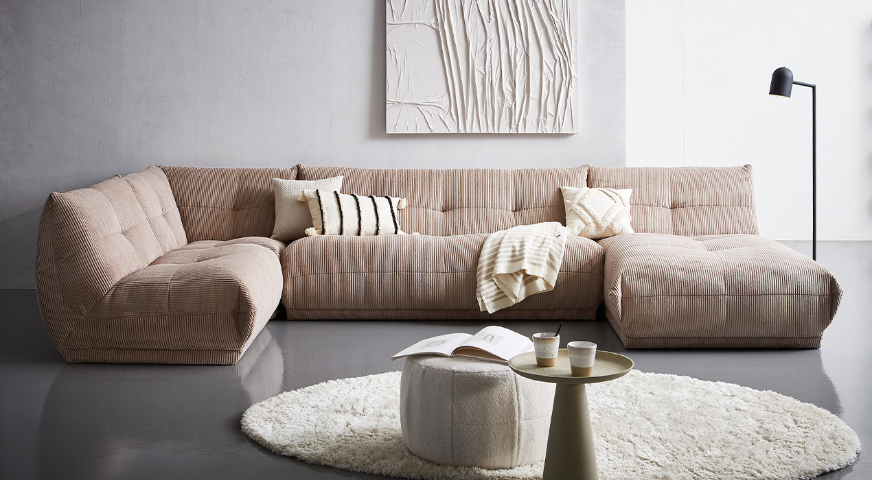 Modernes Wohnzimmer mit einem großen beigen Modulsofa, rundem Teppich und dekorativem Couchtisch. Ergänzt durch stilvolle Kissen und eine dezente Wandgestaltung