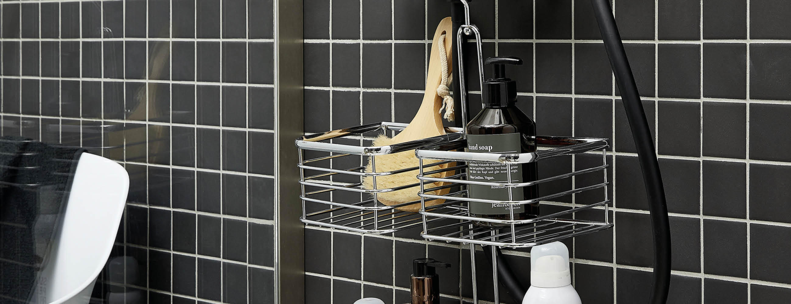 Duschkorb mit Bürste, schwarzer Seifenspenderflasche und weiteren Pflegeprodukten vor schwarz-weißer Fliesenwand
