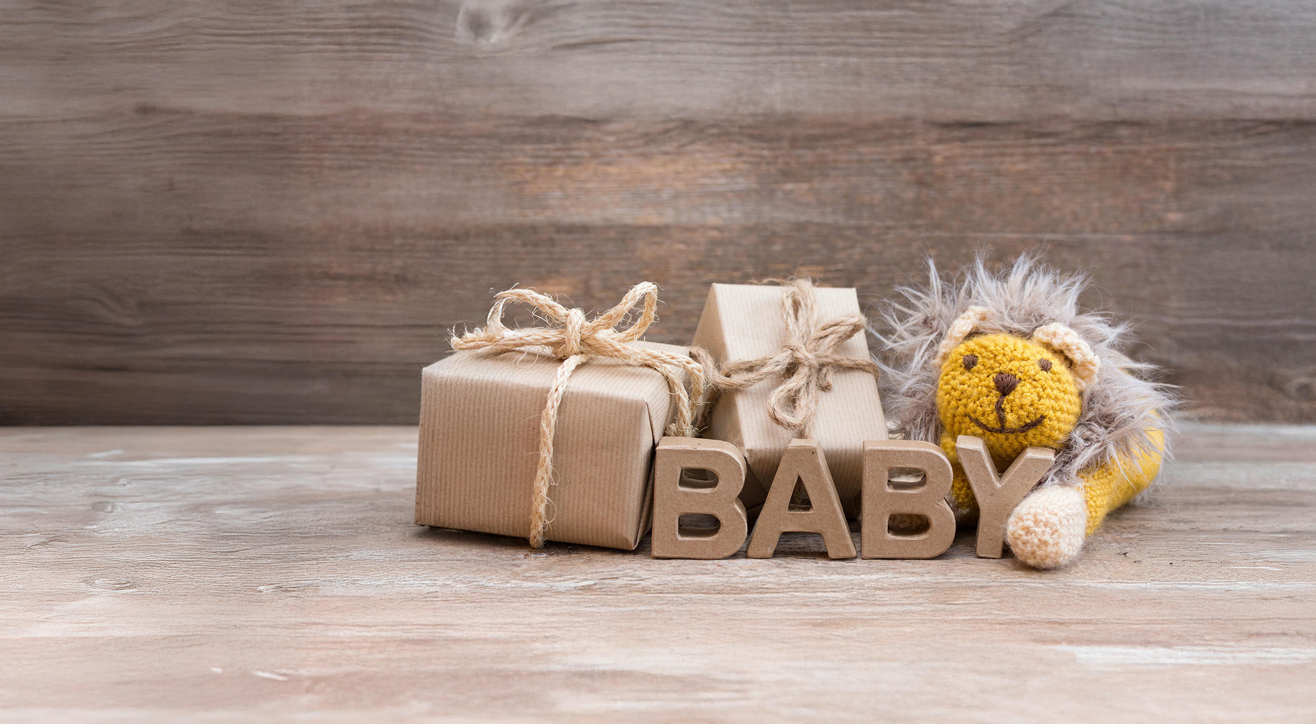 Eine dekorative Anordnung aus zwei eingepackten Geschenken in braunem Papier, einem gehäkelten Löwen‑Plüschtier und großen Buchstaben, die das Wort BABY formen, steht auf einer Holzoberfläche vor einem rustikalen Hintergrund.