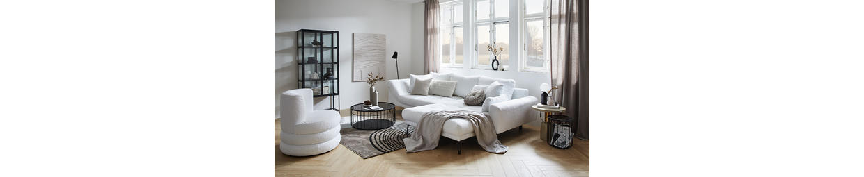 Helles Wohnzimmer mit großem weißen Ecksofa, dekorativen Kissen und Decke, rundem schwarzem Couchtisch mit Vasen und Büchern, hellem Holzboden, Teppich, großen Fenstern mit Vorhängen und abstrakter Wandkunst