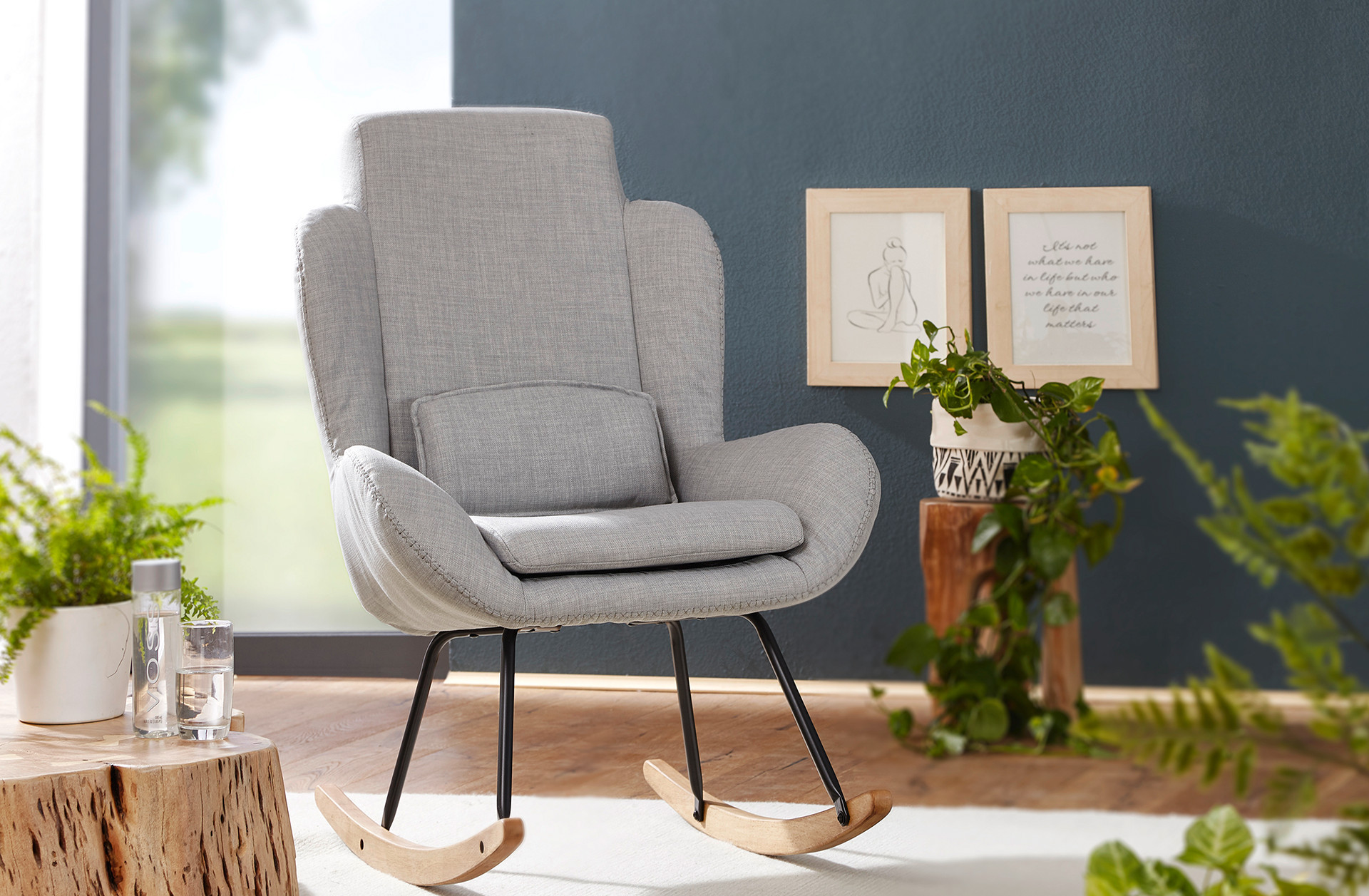 Moderné sivé hojdacie kreslo s čiernymi nohami a drevenými oblúkmi, zelené rastliny v kvetináčoch, drevený stolík s dekoráciami, dva zarámované obrázky na stene