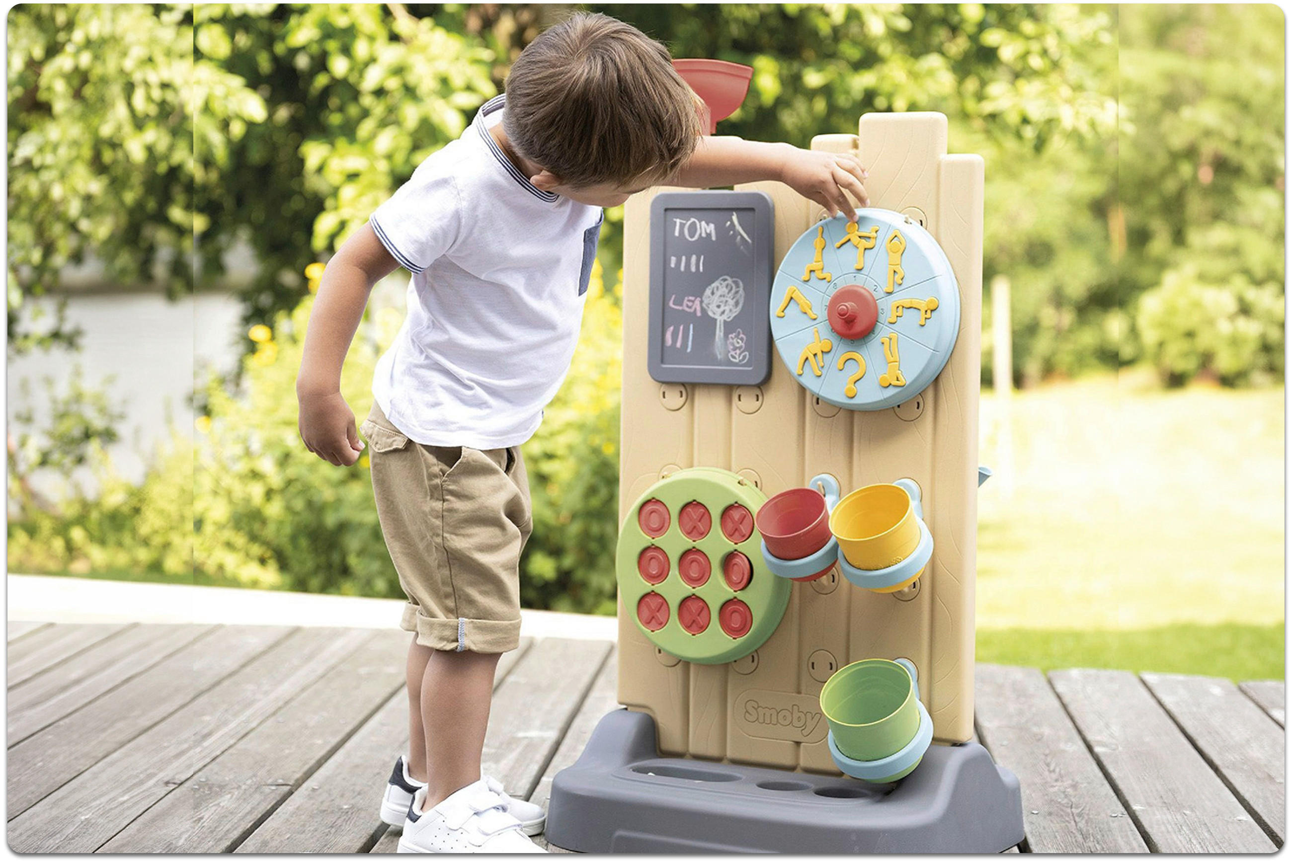 Kleiner Junge steht vor einem Spielgerät mit bunten Bechern, einem blauen Drehrad mit gelben Symbolen und einer kleinen Tafel mit Kreidezeichnungen.