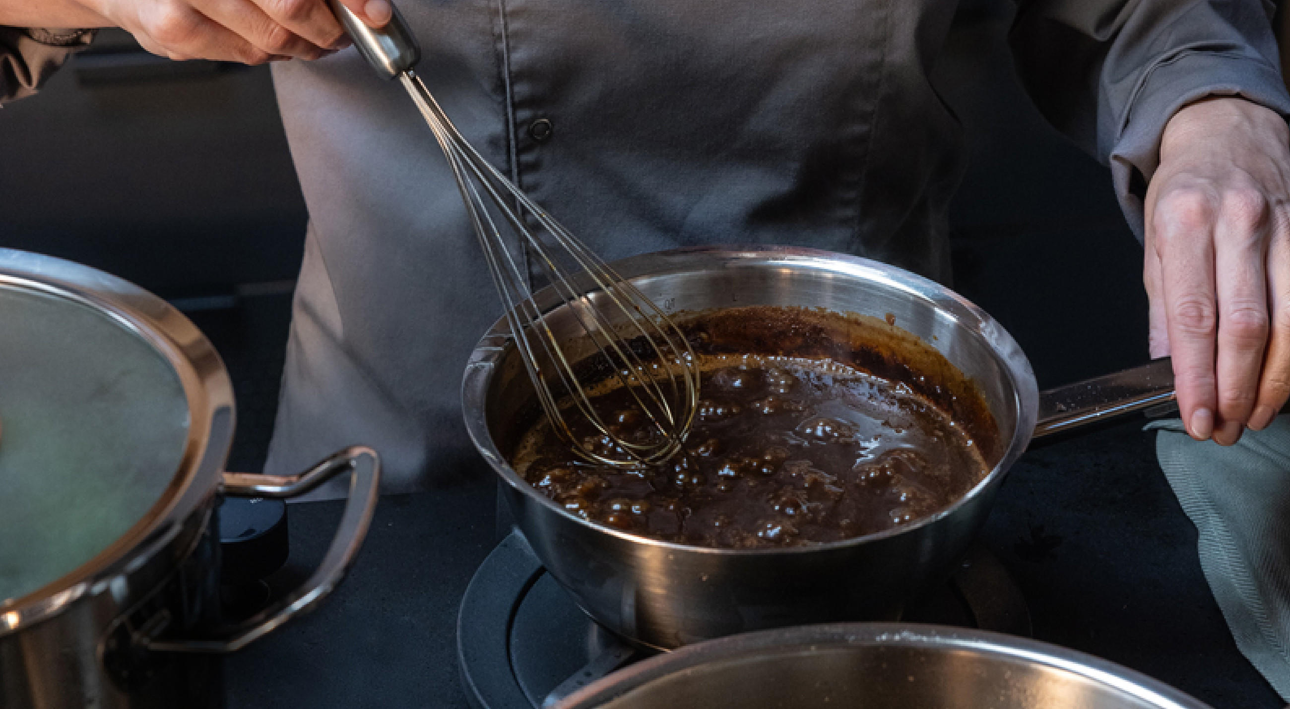 Hände in Kochjacke rühren mit einem Schneebesen von FISSLER in einem Edelstahl‑Topf eine dunkelbraune, blubbernde Sauce; daneben stehen weitere Töpfe auf dem Herd von FISSLER, rechts liegt ein Küchentuch.