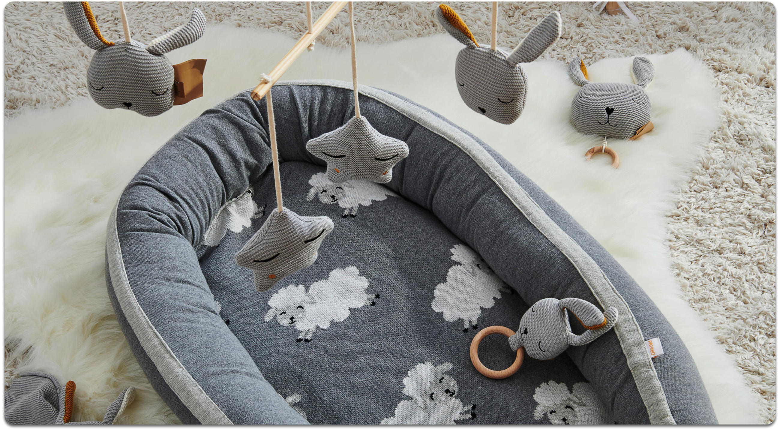 Graues Babynest mit Schafmuster und gepolstertem Rand auf weißem Fellteppich, darüber Mobile mit gestrickten Sternen und Hasenköpfen sowie Rasselring am Rand.