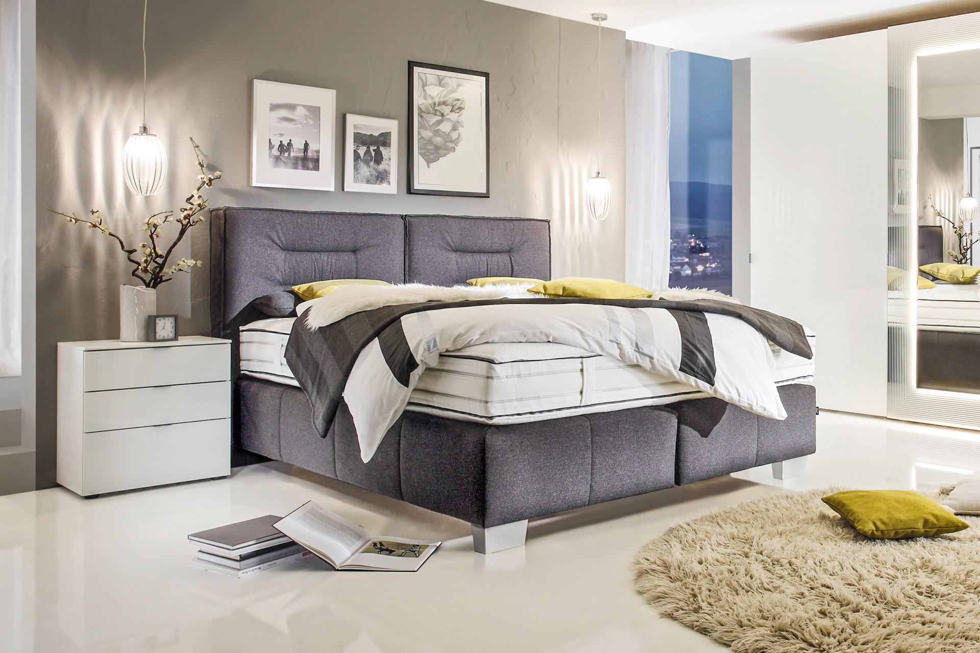 Šedá boxspring posteľ, biely nočný stolík so zásuvkami, závesné lampy, huňatý koberec, obrazy nad posteľou