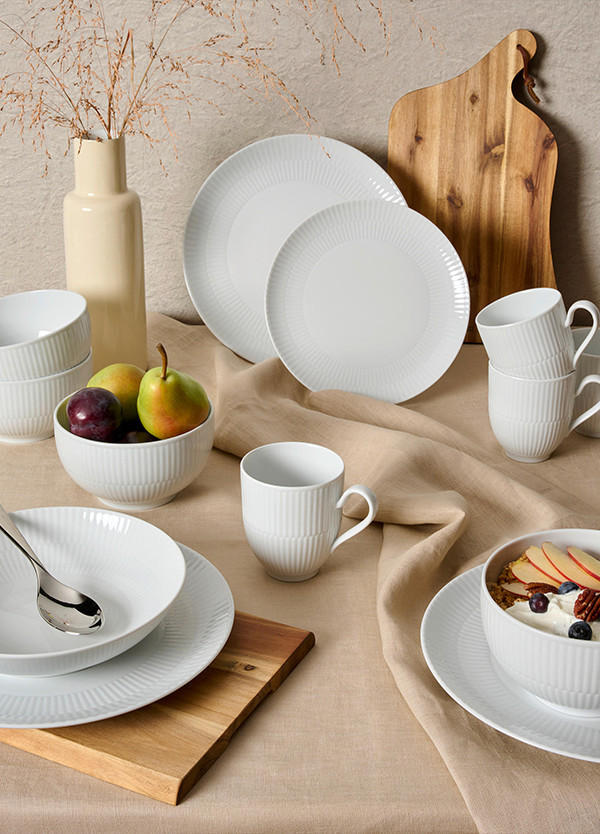 Miza z belo porcelanasto posodo, sadjem in suhim cvetjem na bež prtu.