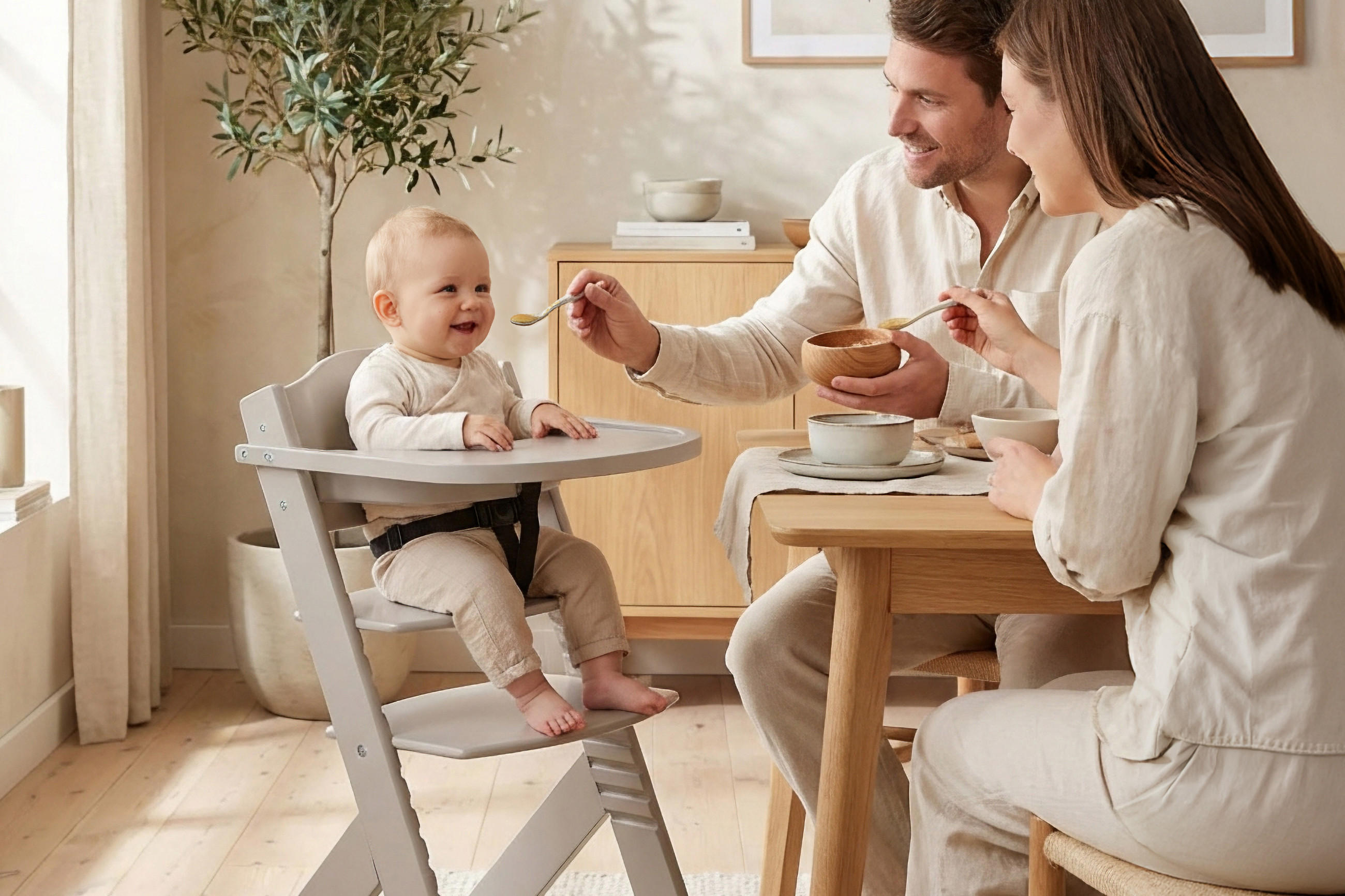 Eltern füttern Baby im Hochstuhl an einem Holztisch in hellem Wohnzimmer.