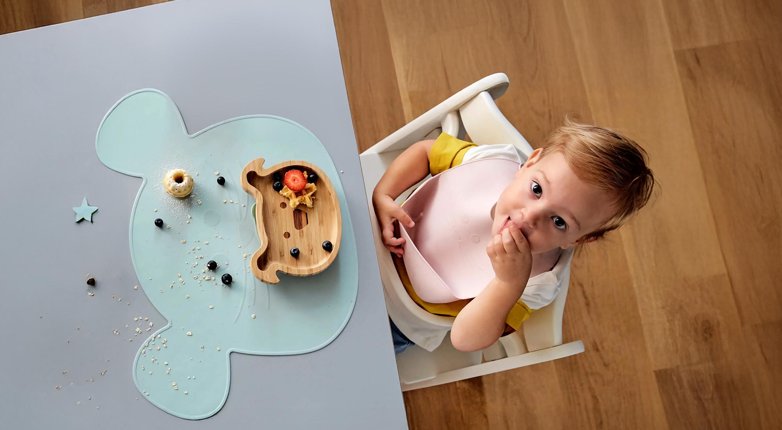 Kleinkind sitzt im Hochstuhl mit Lätzchen und isst, vor sich ein tierförmiges Platzset mit verstreuten Lebensmitteln sowie ein Holz-Teller mit verschiedenen Speisen wie Pasta und Gemüse.
