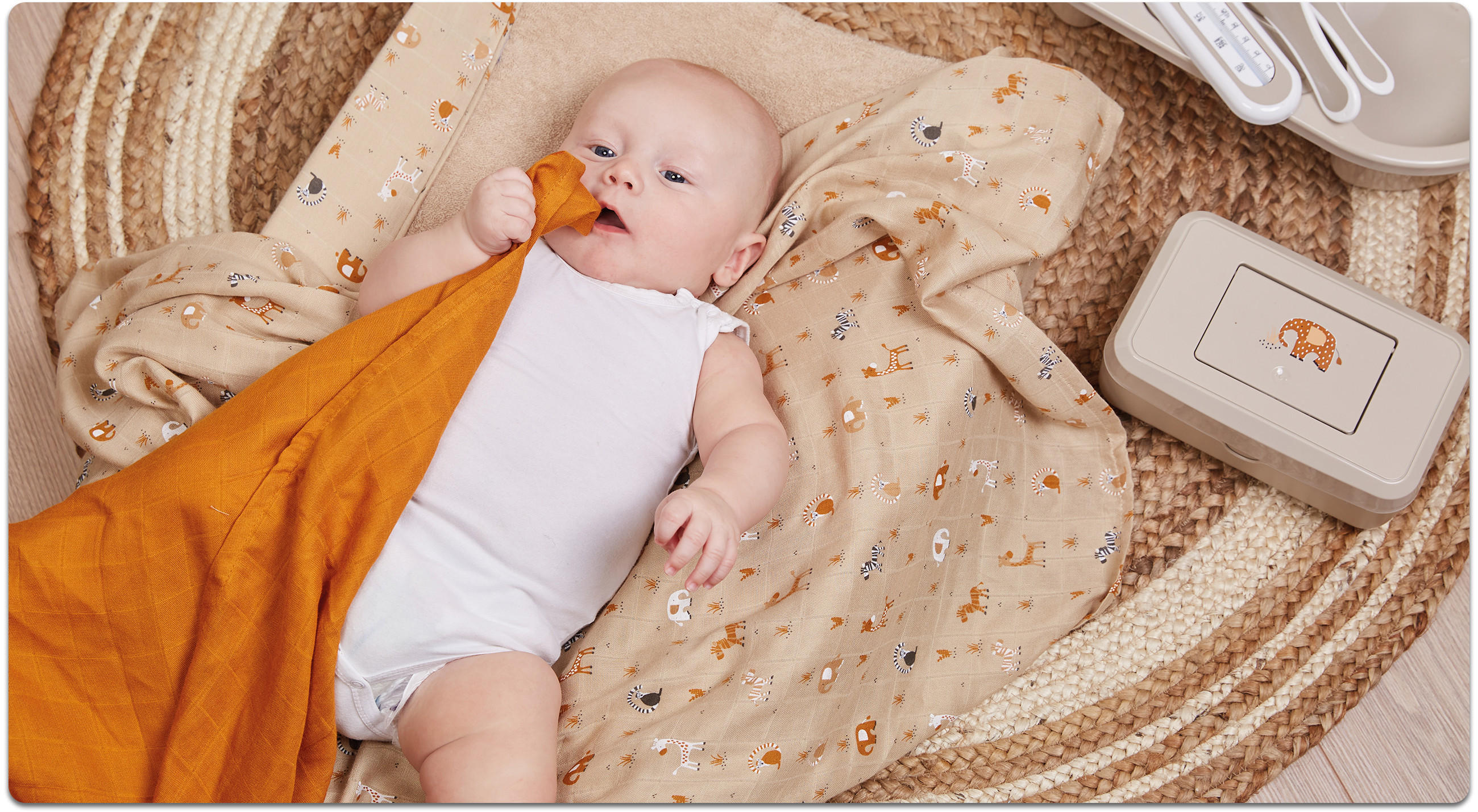 Baby liegt auf gemusterter Decke auf rundem Teppich und kaut auf einem orangefarbenen Tuch; rechts liegen eine Dose und Schuhe.