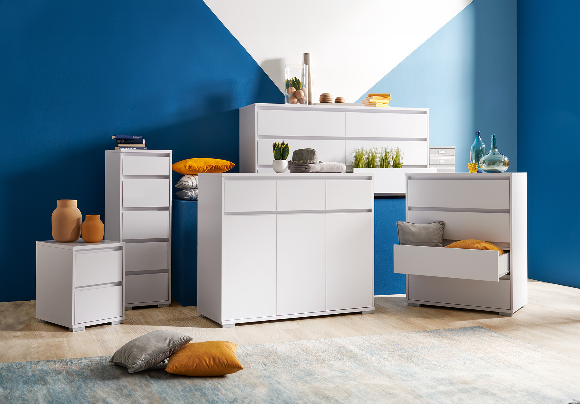 biela zostava drobného nábytku: komody biele, biele komody so zásuvkami, oranžové vankúše, oranžové vázy, modrá stena, drevená podlaha