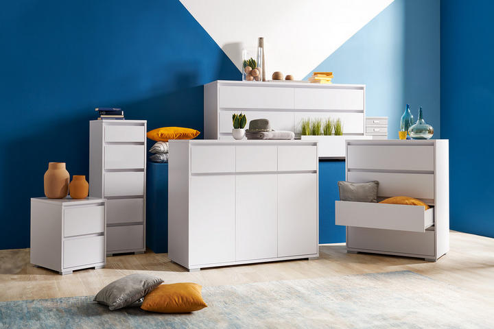 biela zostava drobného nábytku: komody biele, biele komody so zásuvkami, oranžové vankúše, oranžové vázy, modrá stena, drevená podlaha