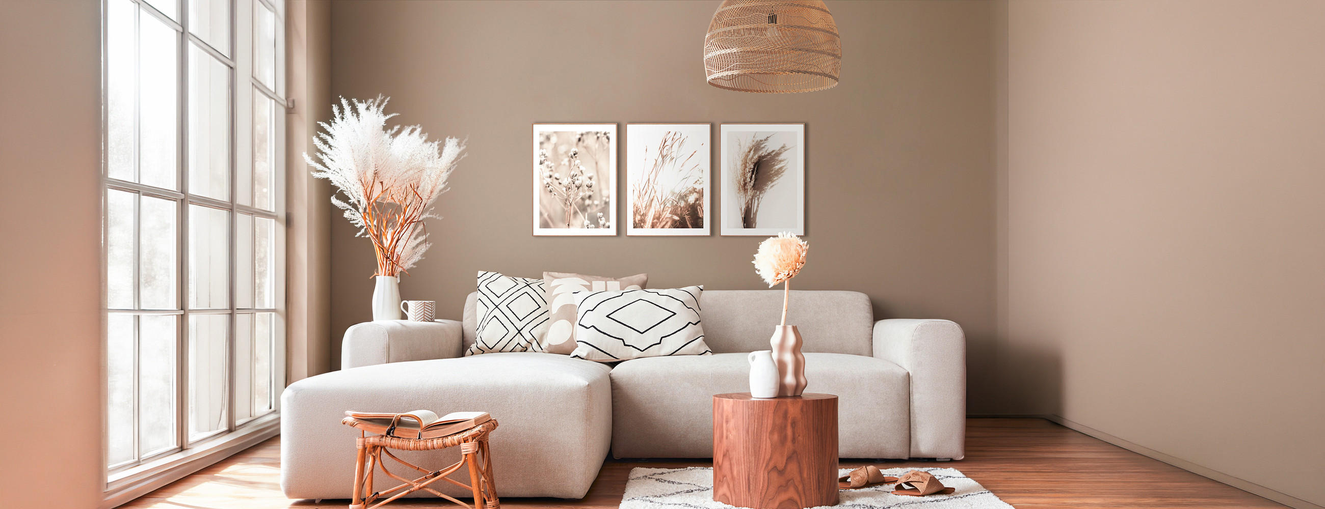 Wohnzimmer mit hellgraßem Sofa, drei gerahmten Pflanzenbildern an der Wand, großem Fenster links und Holzlampe an der Decke.