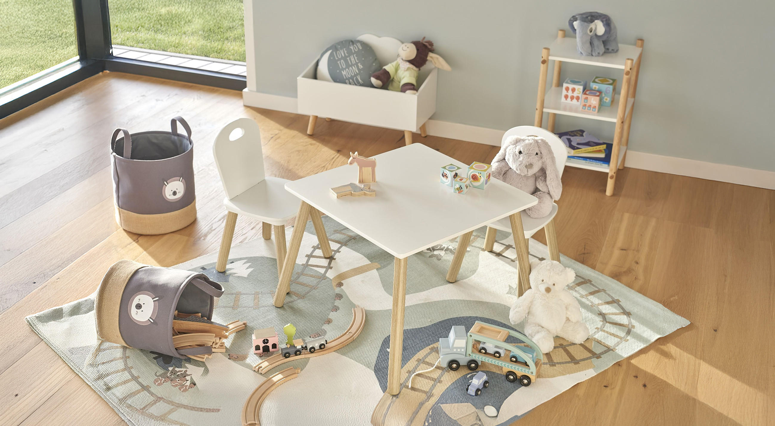Eine helle Spielecke im Wohnzimmer mit kleinem Tisch und zwei Stühlen, einer Spielmatte mit Zugschienen‑Motiv, Stofftieren, Holzspielzeug und Aufbewahrungskörben.
