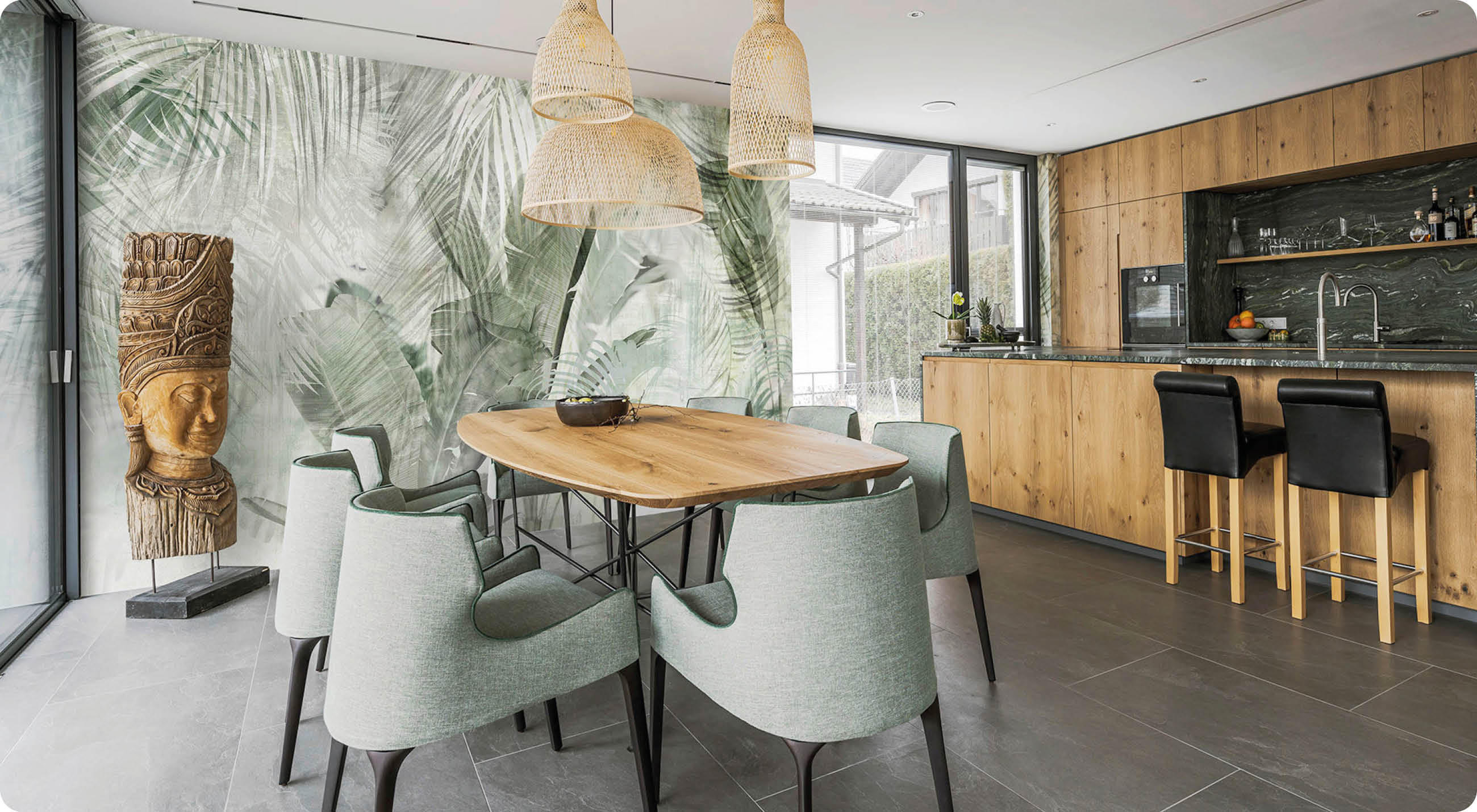 Esszimmer mit großer Wandgestaltung aus Palmenblättern, Holztisch mit grauen Stühlen, Holzvertäfelung und moderner Küchenzeile mit Barhockern