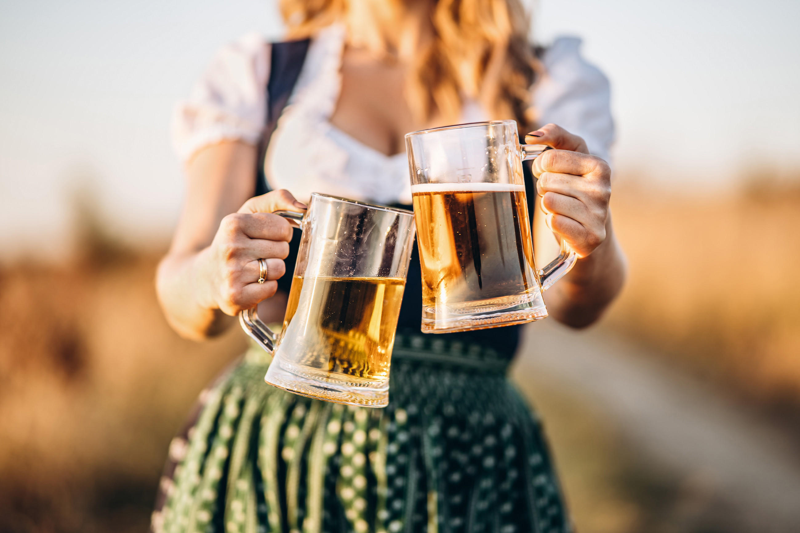 Person in Dirndl stößt beim Oktoberfest mit zwei Bierkrügen an – helle Biere mit Schaum, draußen bei Tageslicht.