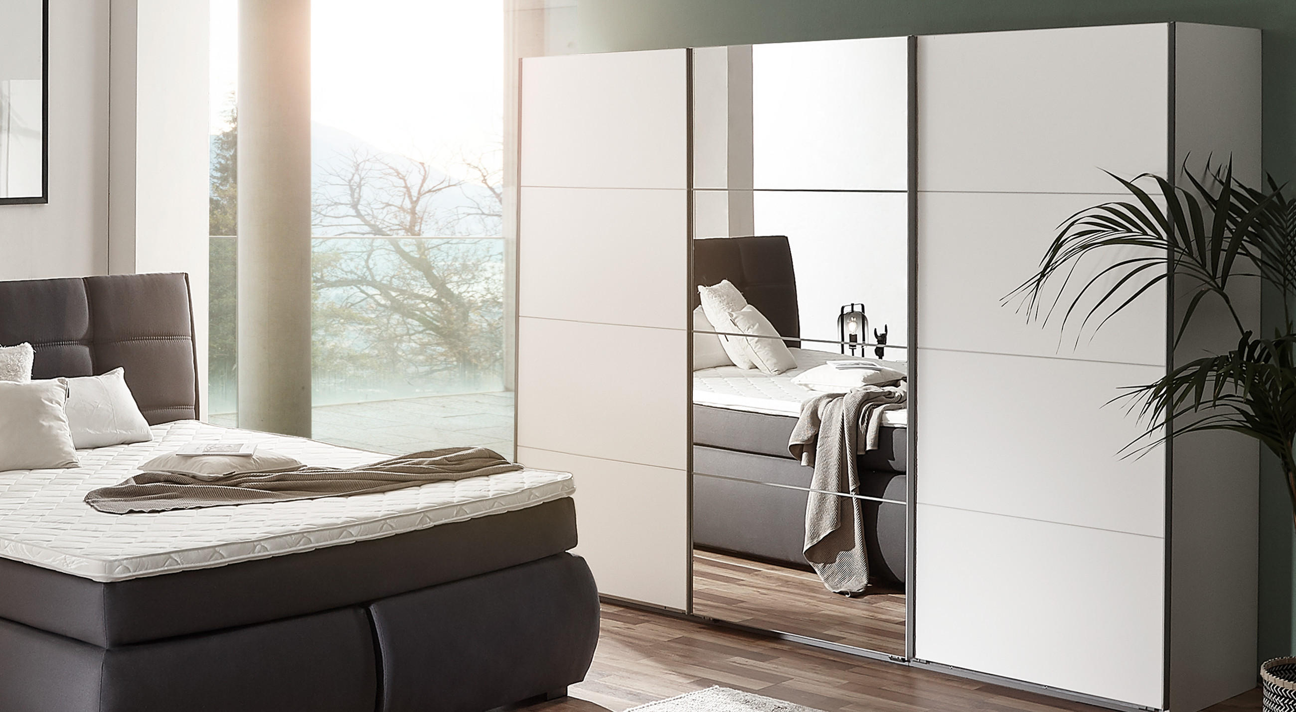 Modernes Schlafzimmer mit dunkelgrauem Polster‑Boxspringbett, weißer Matratzenauflage und schlichtem Plaid; rechts großer weißer Schwebeschrank mit Spiegelfront, Parkettboden und hoher Zimmerpflanze