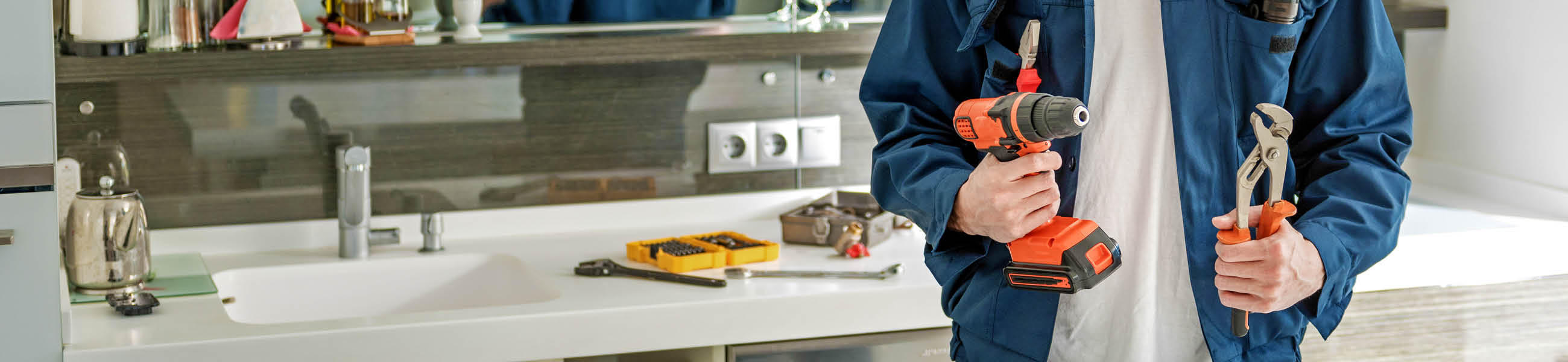 Mann in blauer Arbeitsjacke hält eine Bohrmaschine und eine Zange in der Hand, vor einer modernen Kücheneinrichtung mit Werkzeugen im Hintergrund.