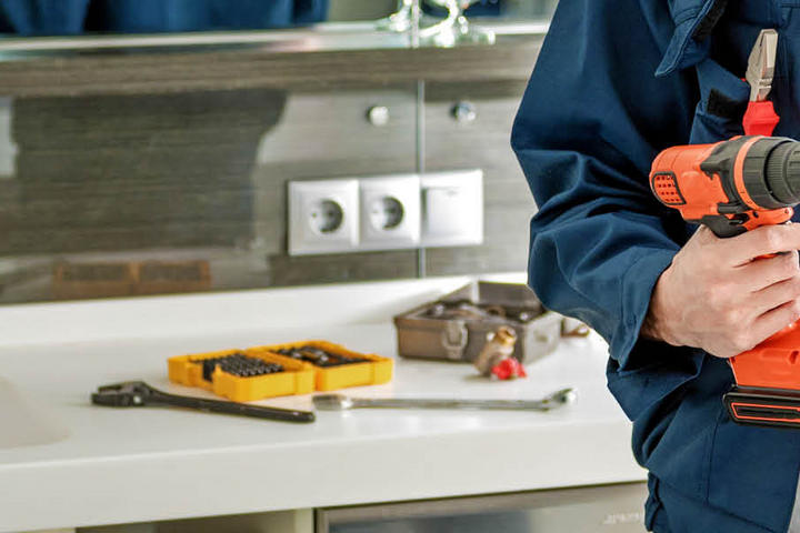 Heimwerker in blauer Jacke hält eine Bohrmaschine und eine Zange in der Hand, vor einer modernen Kücheneinrichtung mit Werkzeugen im Hintergrund.