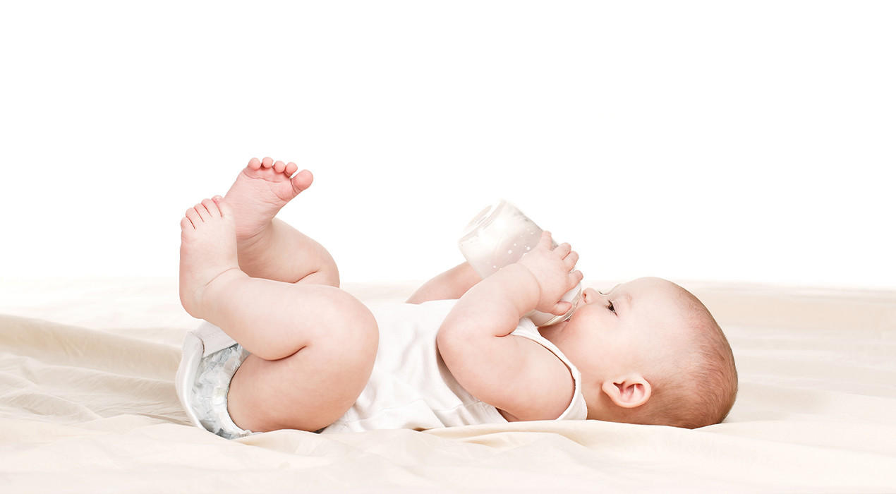 Baby mit Milchflasche auf dem Bett in weißem Strampler