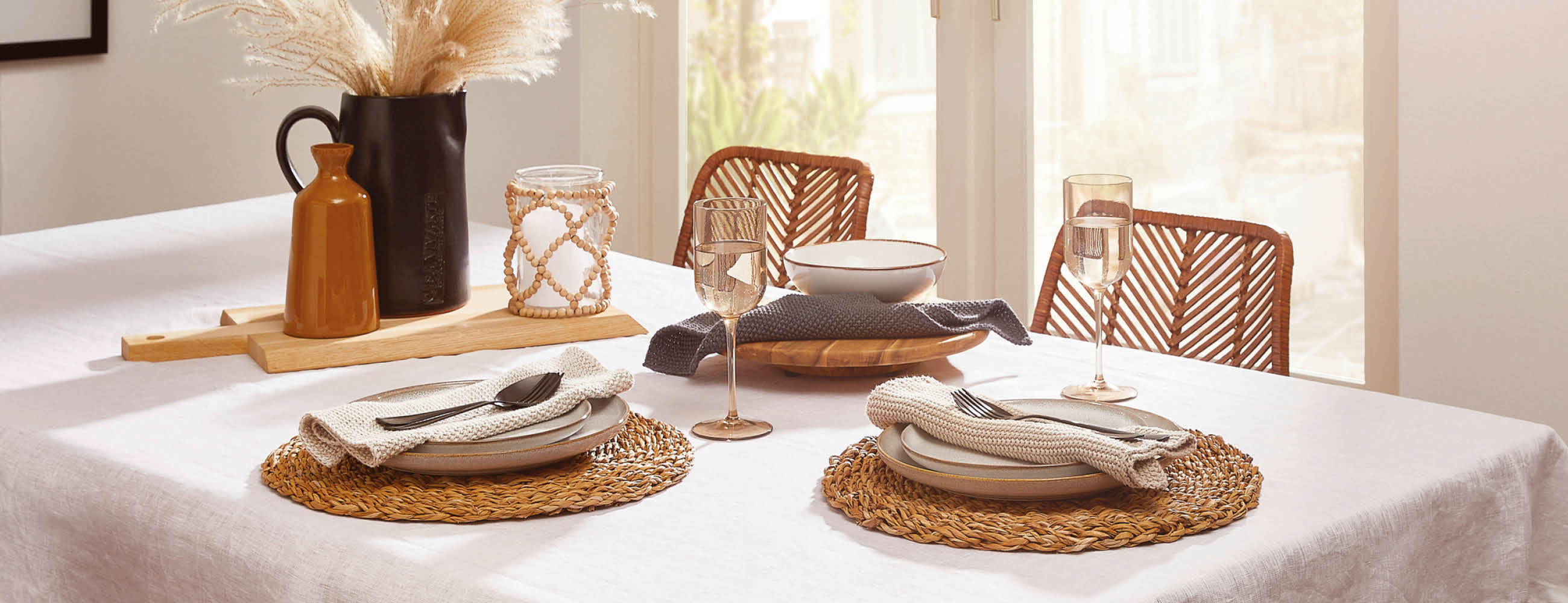 Esszimmertisch mit weißer Tischdecke, zwei gedeckten Plätzen mit geflochtenen Platzsets, Besteck, Gläsern und dekorativen Vasen im Hintergrund