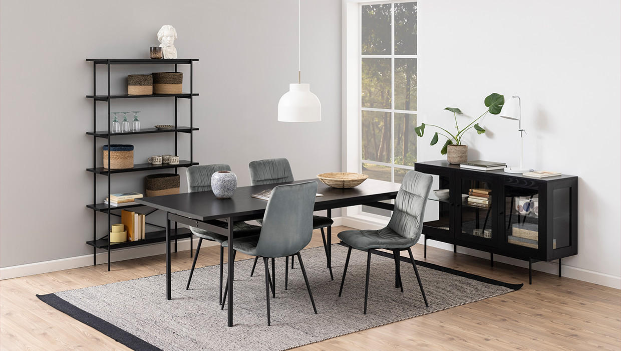 Moderná jedáleň, striedmy vzhľad, tmavá skrinka na nožičkách, presklená skrinka, čierne police, čierny stôl, sivé stoličky, sivý koberec s čiernym lemom, drevená podlaha