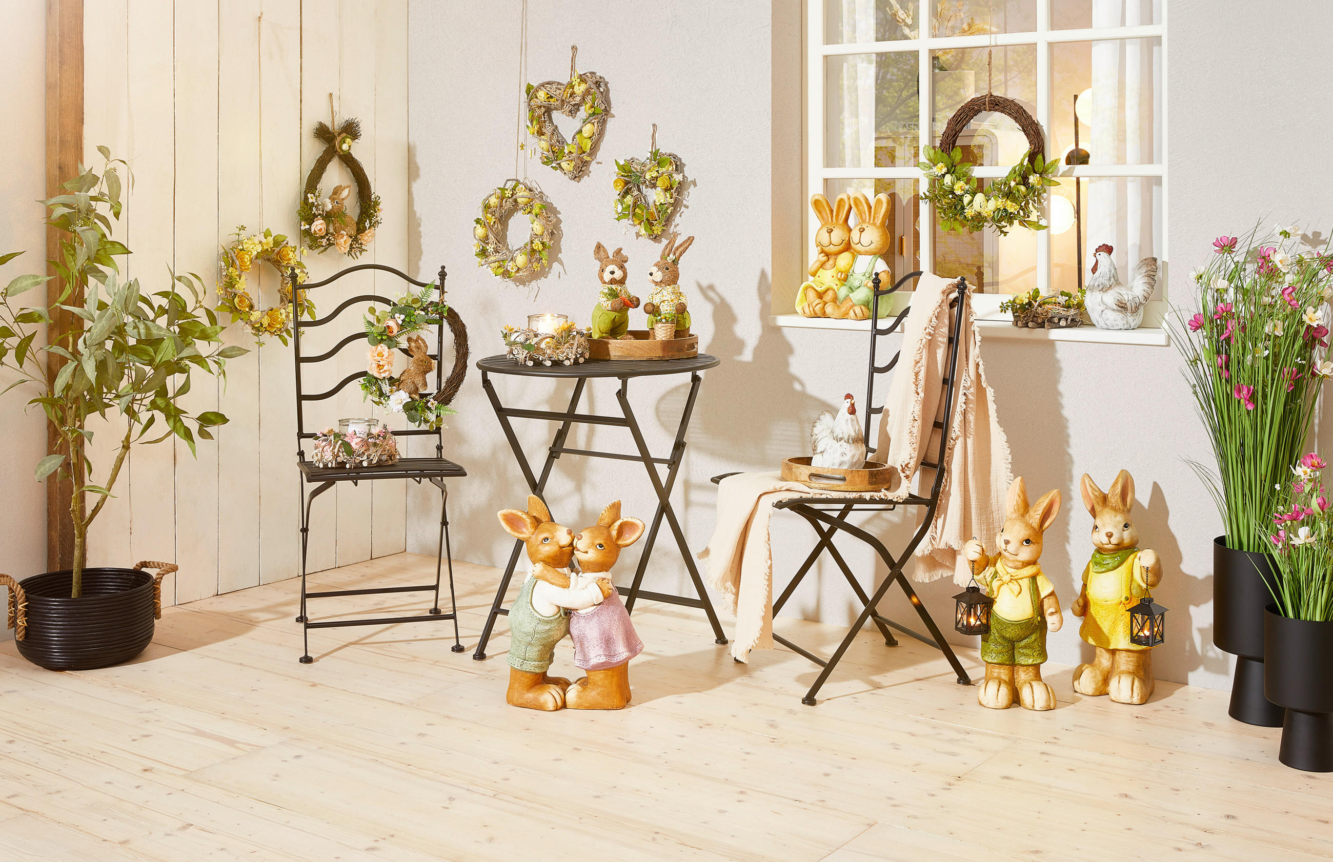 Osterdekoration mit Hasenfiguren, Blumenkränzen an der Wand, zwei Stühlen und einem kleinen Tisch in einem hellen Raum mit Holzboden.