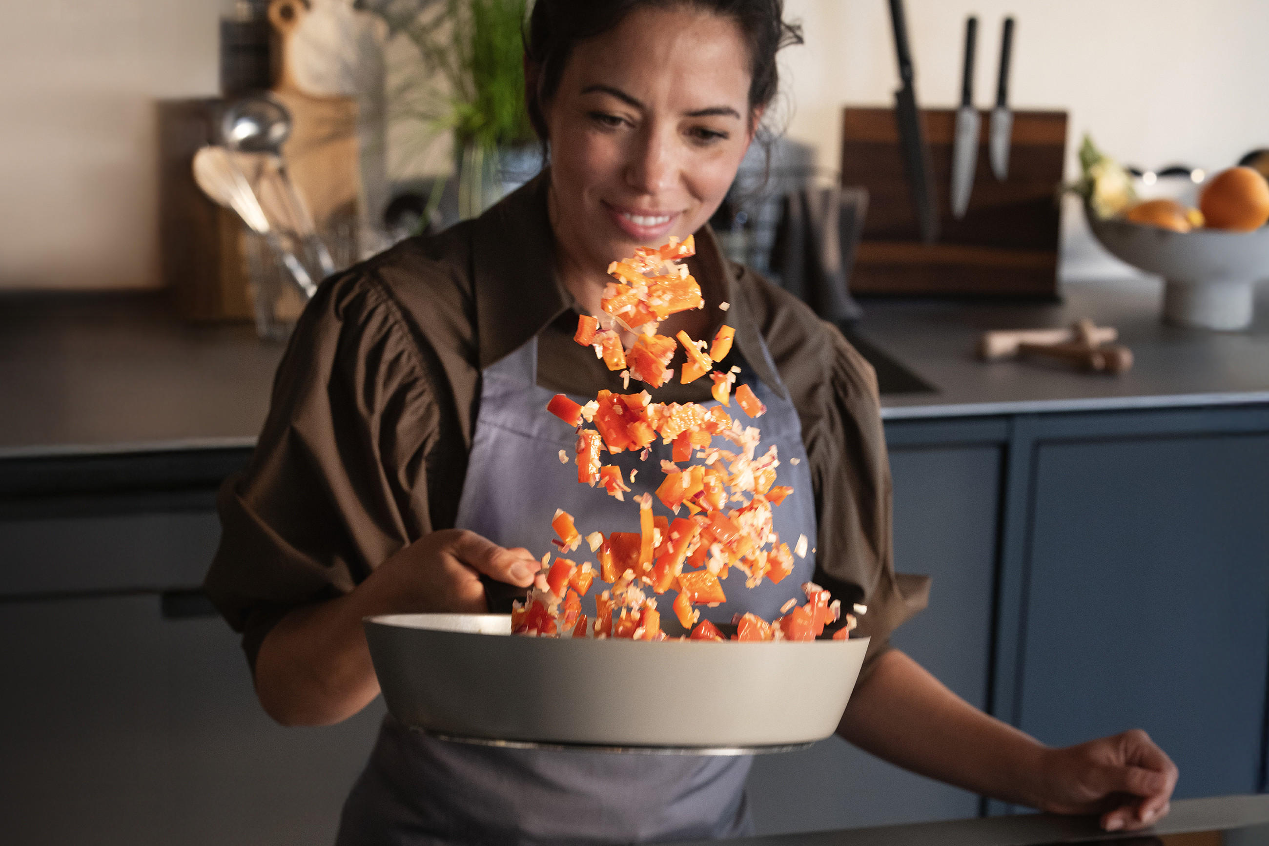 Frau in Schürze schwenkt eine helle Fissler Ceratal® Evo Pfanne, wobei gewürfeltes Gemüse in einem Bogen durch die Luft fliegt; im Hintergrund eine moderne Küche mit Messern und Obstschale.