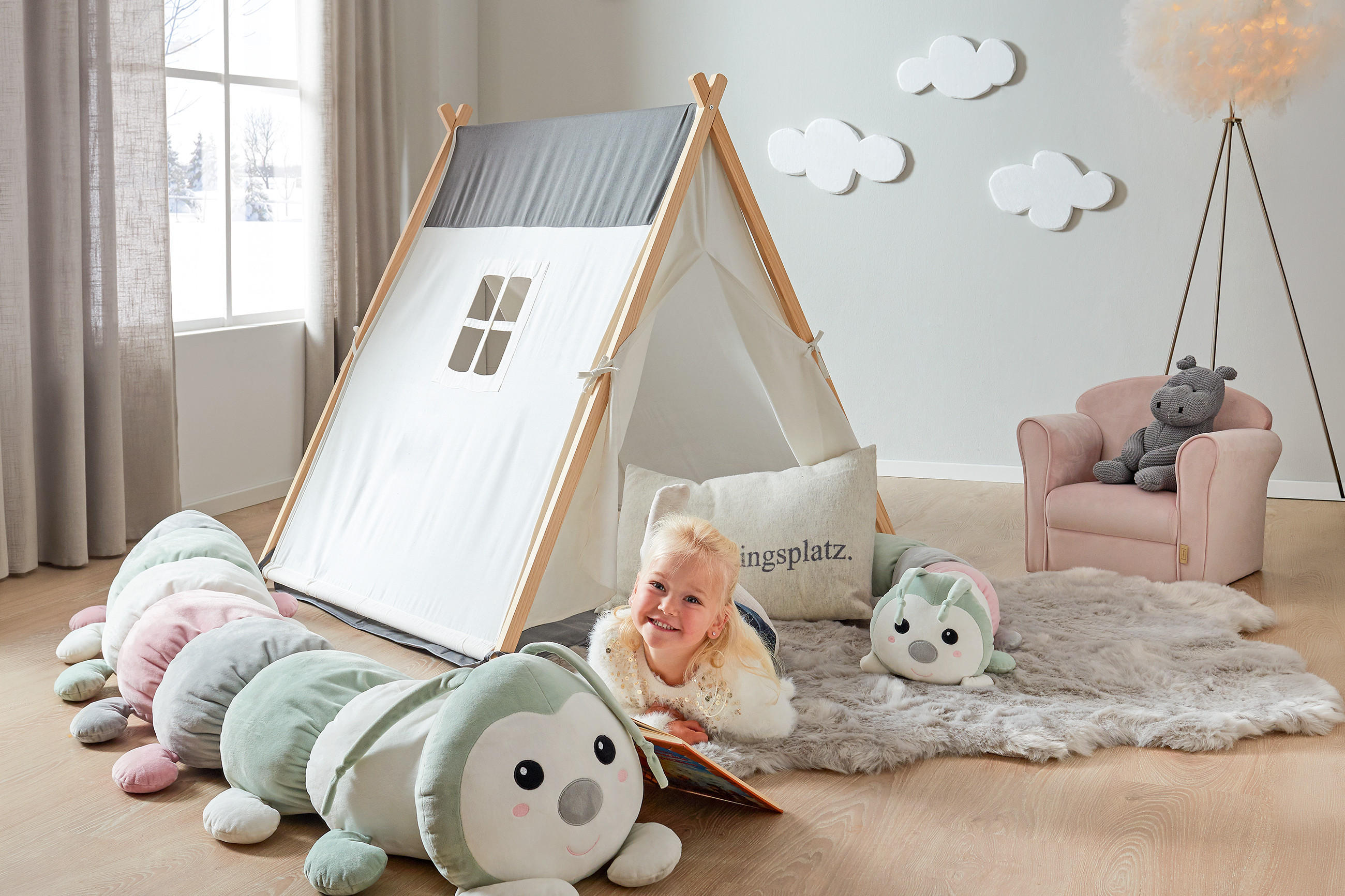 Kind liegt auf dem Boden vor einem Zeltspielhaus mit Fenster und liest ein Buch neben einem großen, weichen Raupenkuscheltier in einem hellen Kinderzimmer mit Teppich, Sessel und Wolkenwanddekoration.