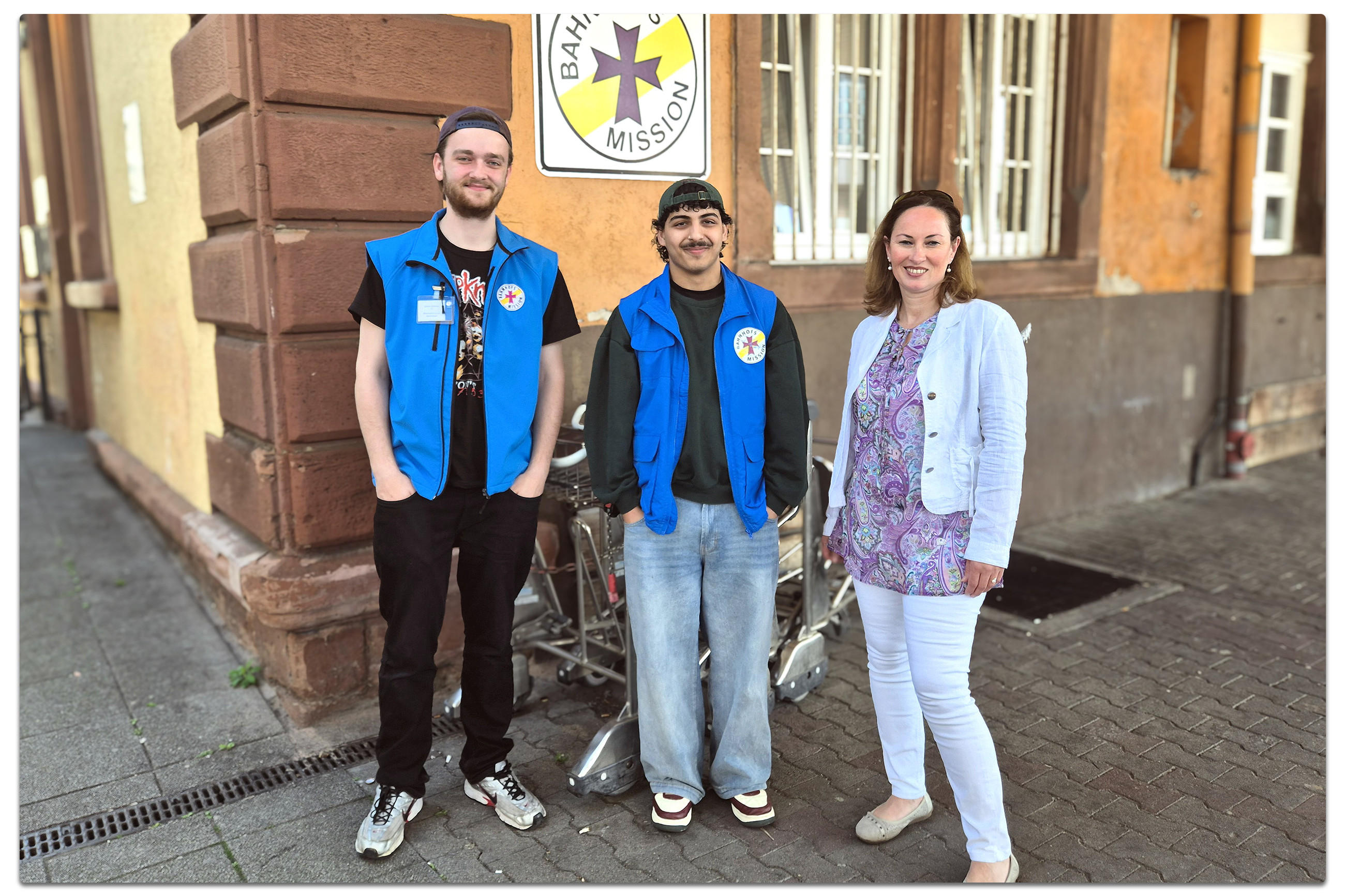 Drei Personen stehen vor einem Gebäude und lächeln in die Kamera.