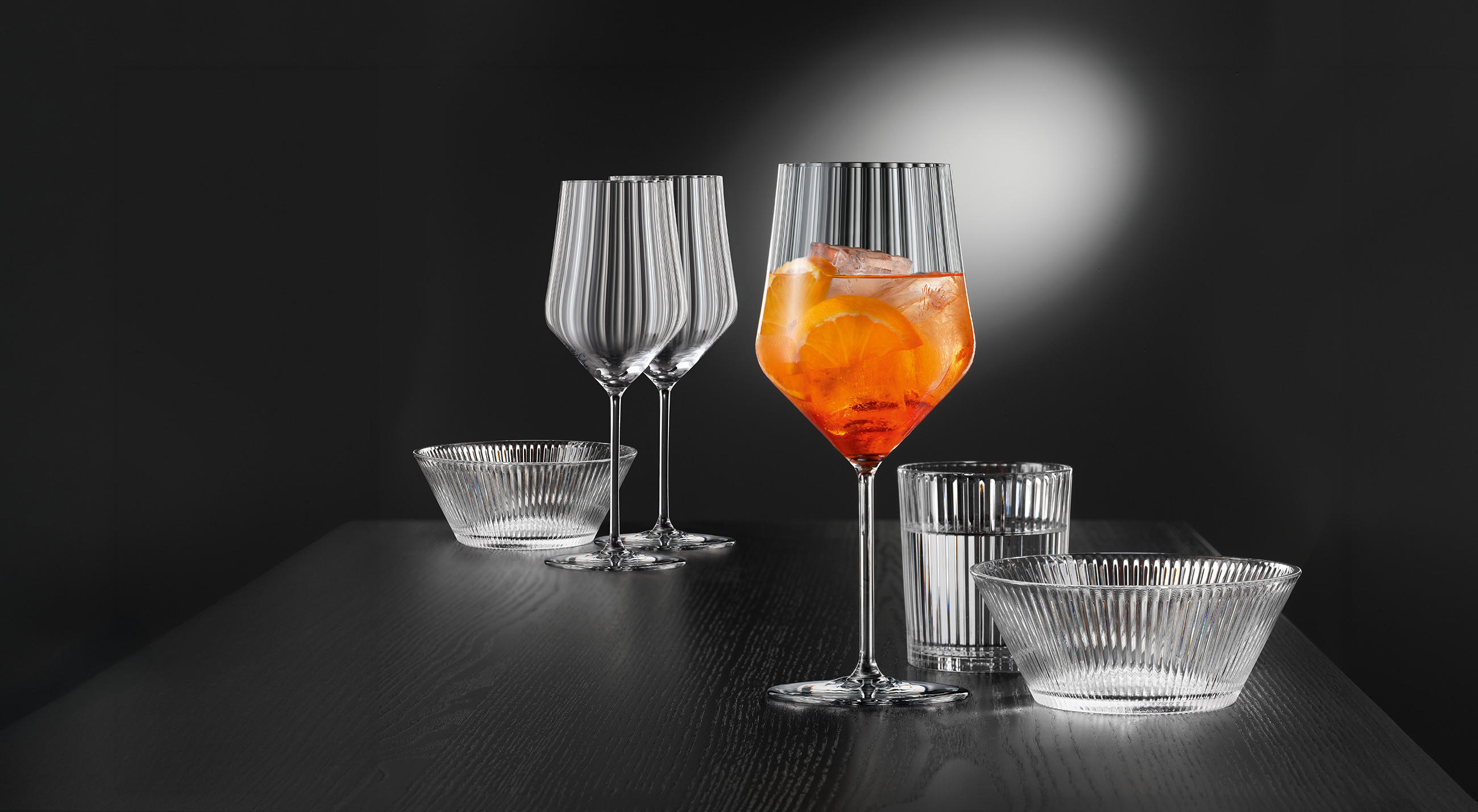 NACHTMANN Gläser-Arrangement auf dunklem Tisch: zwei geriffelte Weingläser, ein Longdrinkglas und Schalen; rechts ein großes Stielglas mit orangefarbenem Cocktail, Eiswürfeln und Orangenscheibe vor dunklem Hintergrund.