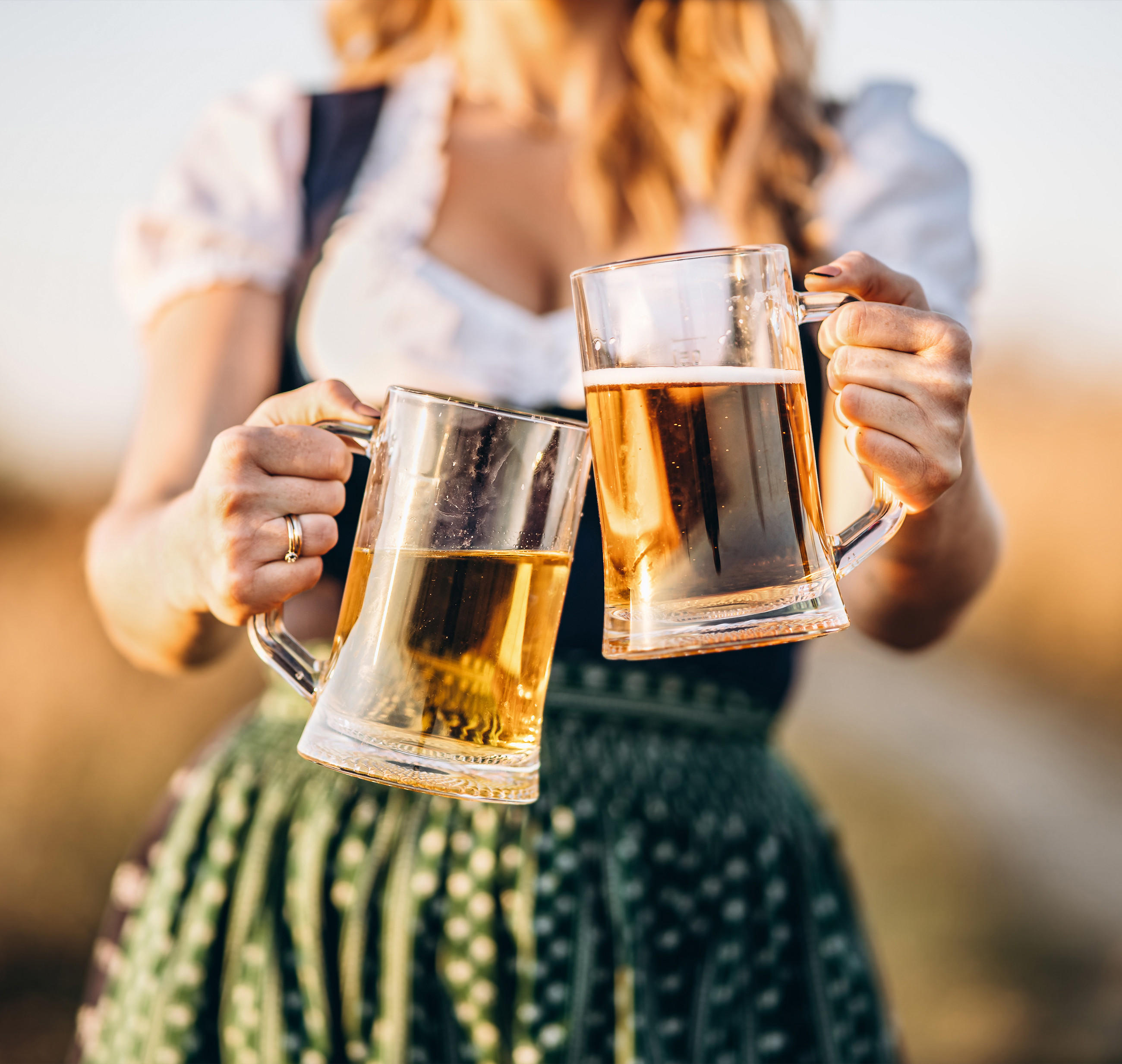 Person in Dirndl stößt beim Oktoberfest mit zwei Bierkrügen an – helle Biere mit Schaum, draußen bei Tageslicht.