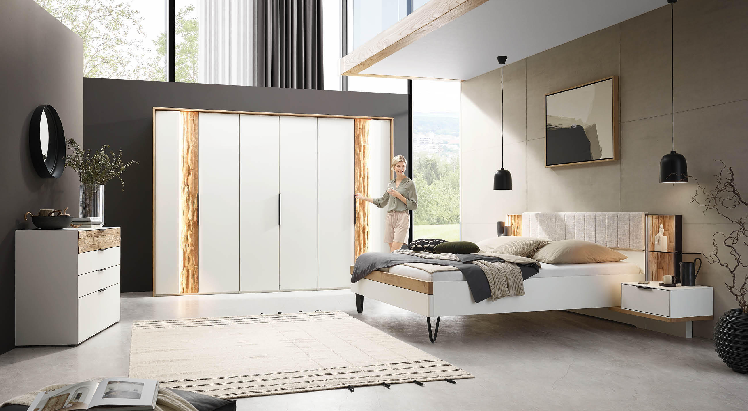 Modern gestaltetes Schlafzimmer in Weiß mit sichtbaren Holzelementen, auch am Bett. Ausgestattet mit einem Kleiderschrank, einer Kommode sowie einem Bett mit integriertem Licht; das Nachttischchen ist direkt am Bett angebracht.