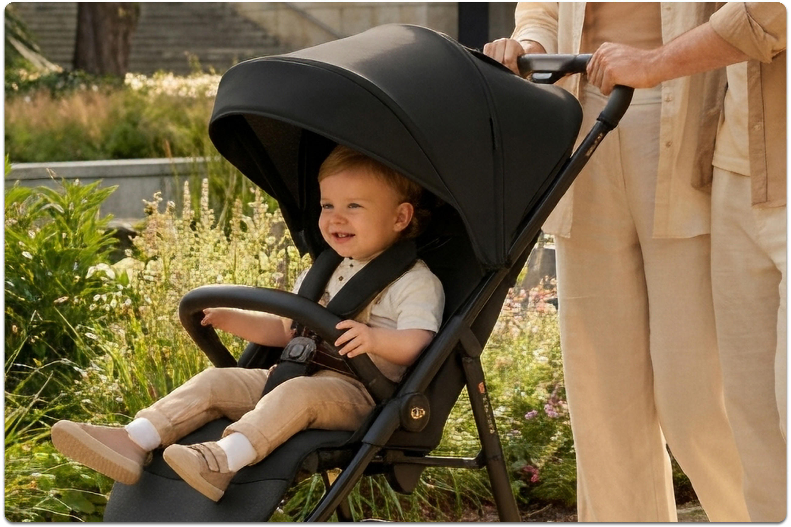 Baby sitzt in schwarzem Kinderwagen und lacht. Die Hände der Eltern sind am Griff des Kinderwagens und sie stehen hinter dem Kinderwagen. Im Hintergrund sind grüne Pflanzen.