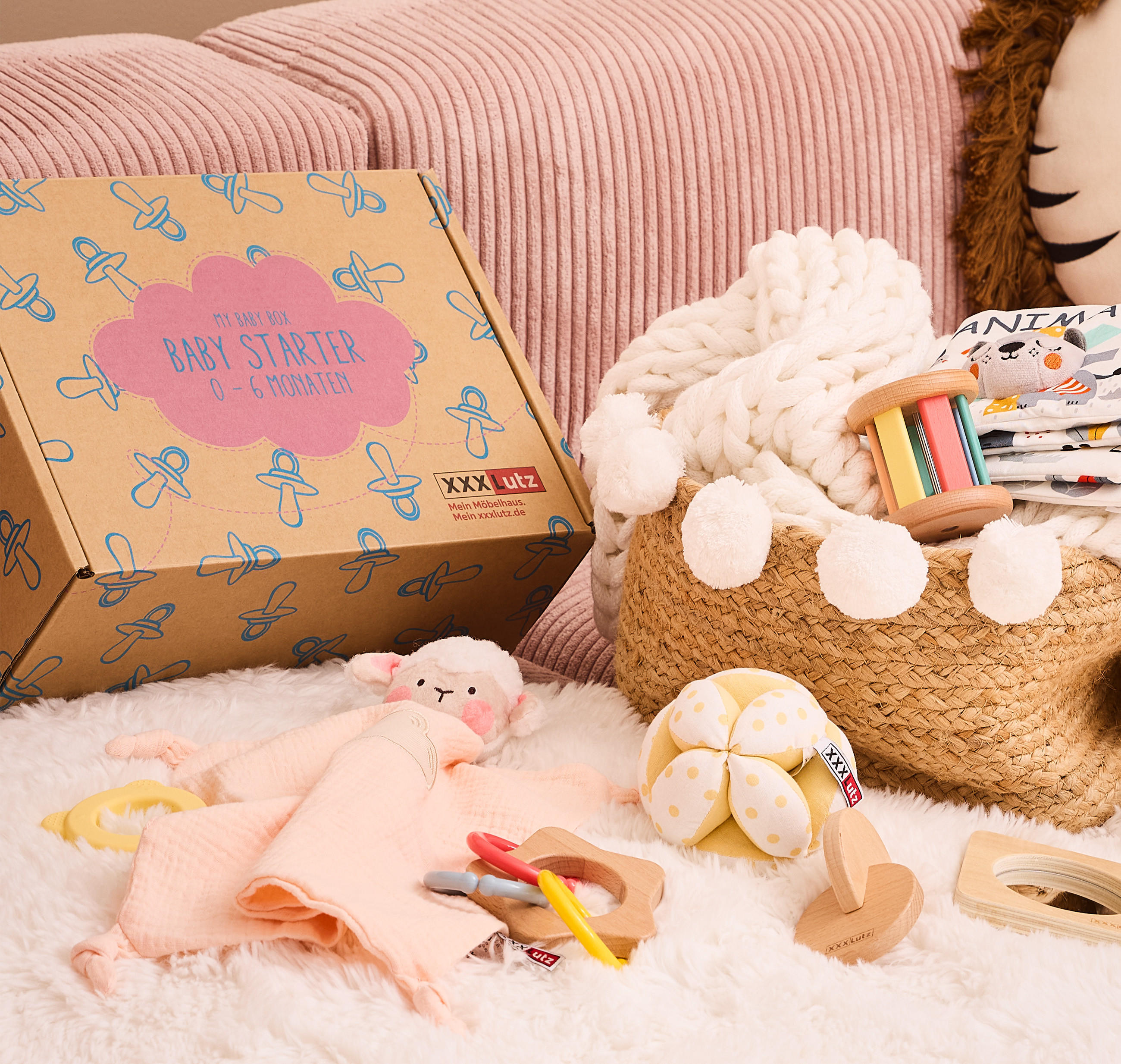 Baby-Starter-Box für 0–6 Monate von XXXLutz neben einem Korb mit weißer Strickdecke, Holzrassel und Stoffbüchern mit Tiermotiven. Im Hintergrund rosa Kissen und ein Dekokissen mit Tiergesicht